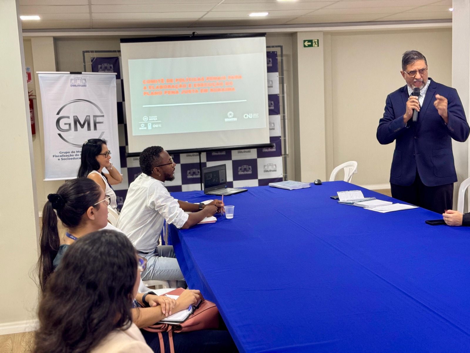 Imagem colorida contém o assessor especial do GMF, em pé ao lado da mesa e reunião, realizando apresentação em slide, durante a primeira reunião do comitê de políticas penais de Roraima. Ao lado oposto da mesa está quatro integrantes do colegiado. Atras deles encontram-se o powerpoint e ao lado um banner do GMF/TJRR. Imagem colorida contém o assessor especial do GMF, em pé ao lado da mesa e reunião, realizando apresentação em slide, durante a primeira reunião do comitê de políticas penais de Roraima. Ao lado oposto da mesa está quatro integrantes do colegiado. Atras deles encontram-se o powerpoint e ao lado um banner do GMF/TJRR.