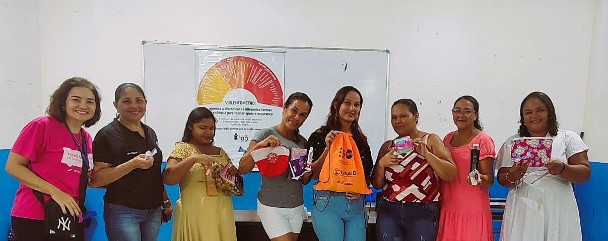 A imagem mostra um grupo de oito mulheres em pé, posando para uma foto. As mulheres na foto parecem estar segurando pequenos sacos ou nécessaires de tecido com diferentes estampas e cores. A imagem mostra um grupo de oito mulheres em pé, posando para uma foto. As mulheres na foto parecem estar segurando pequenos sacos ou nécessaires de tecido com diferentes estampas e cores.