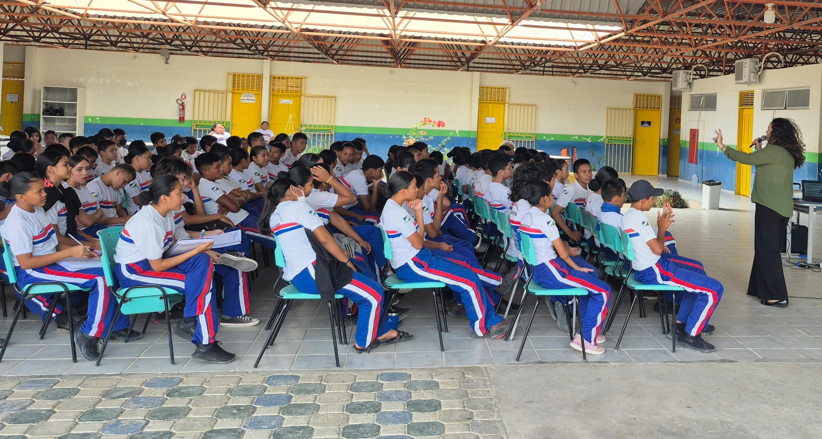 A imagem mostra um grande de estudantes, sentados em um espaço amplo e aberto, que parece ser um pátio coberto ou uma área comum de uma escola. A coordenadora da Justiça Comunitária, Marcelle Wottrich, está a frente, discursando. A imagem mostra um grande de estudantes, sentados em um espaço amplo e aberto, que parece ser um pátio coberto ou uma área comum de uma escola. A coordenadora da Justiça Comunitária, Marcelle Wottrich, está a frente, discursando.