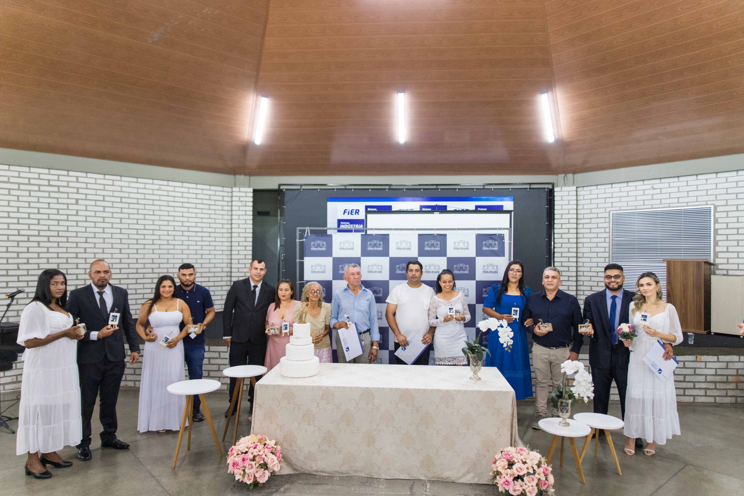  Imagem colorida mostra 14 pessoas — os sete casais que oficializaram matrimônio no primeiro Casamento Coletivo da Corregedoria-Geral de Justiça de Roraima. Ao fundo, vê-se um painel backdrop do Judiciário Roraimense e, à frente, uma mesa com um bolo de casamento.