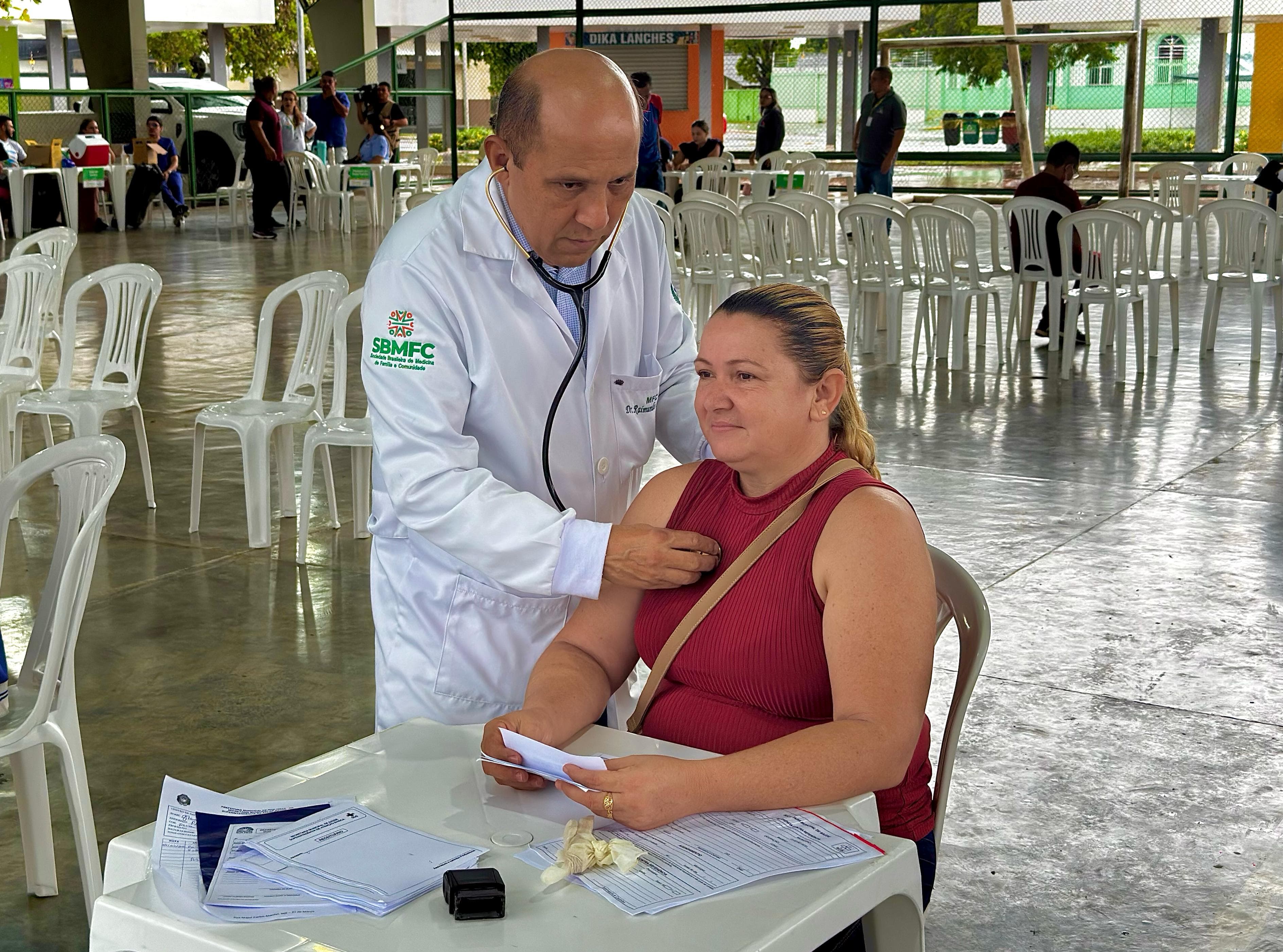 oto colorida mostrando um médico, examinando uma paciente sentada. O médico está usando um estetoscópio para auscultar o peito da mulher. A paciente está vestindo uma blusa vermelha e tem uma bolsa a tiracolo. Ela parece estar segurando alguns papéis em suas mãos.oto colorida mostrando um médico, examinando uma paciente sentada. O médico está usando um estetoscópio para auscultar o peito da mulher. A paciente está vestindo uma blusa vermelha e tem uma bolsa a tiracolo. Ela parece estar segurando alguns papéis em suas mãos.