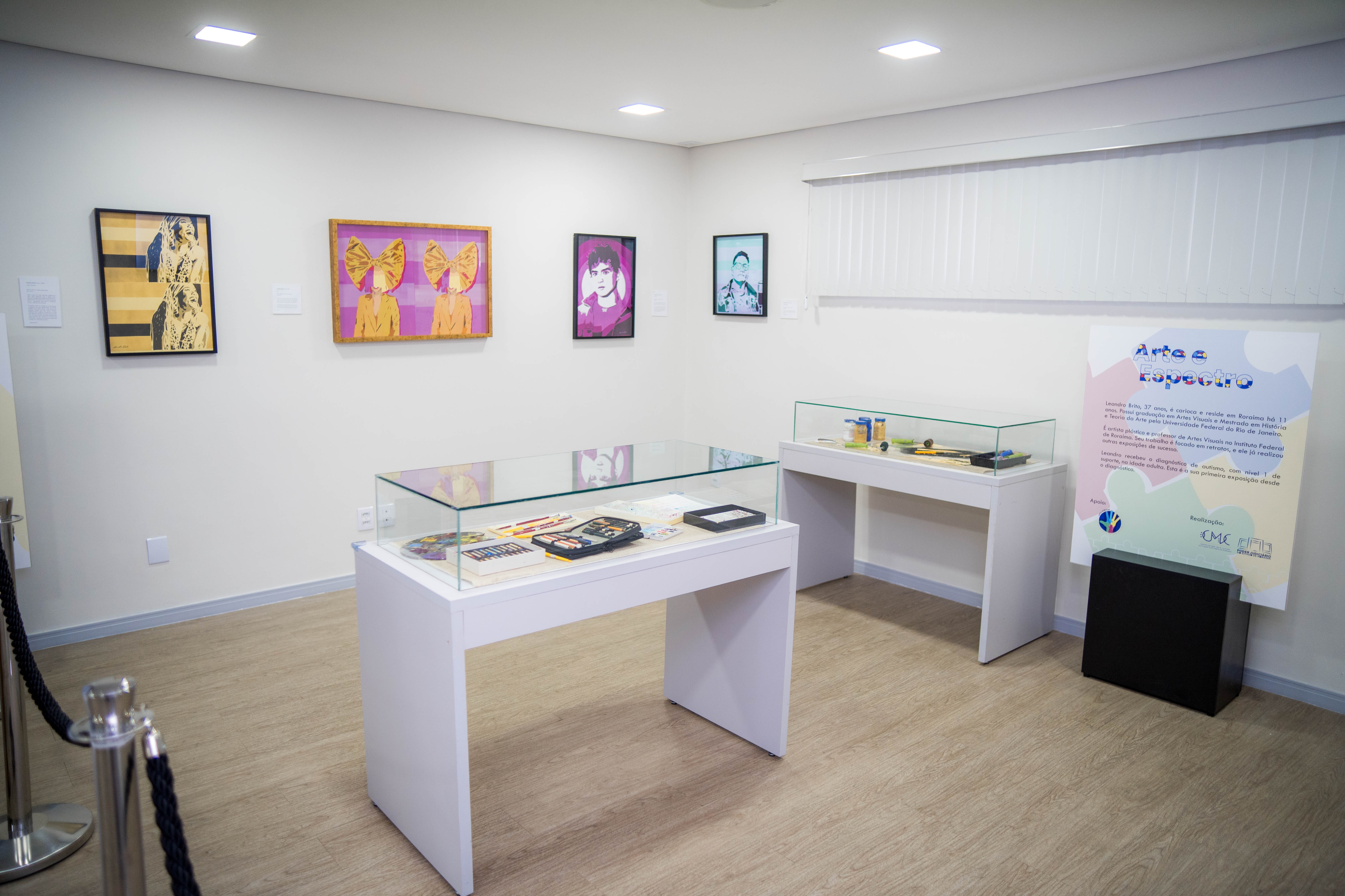 A imagem mostra o interior de uma sala de exposição. As paredes são brancas e quadros estão pindurados na parede. No centro uma vitrine, com diversos materiais de arte, incluindo uma paleta de pintura colorida com tinta seca, pincéis de diferentes tamanhos, estojos com lápis de cor e outros materiais de desenho.
