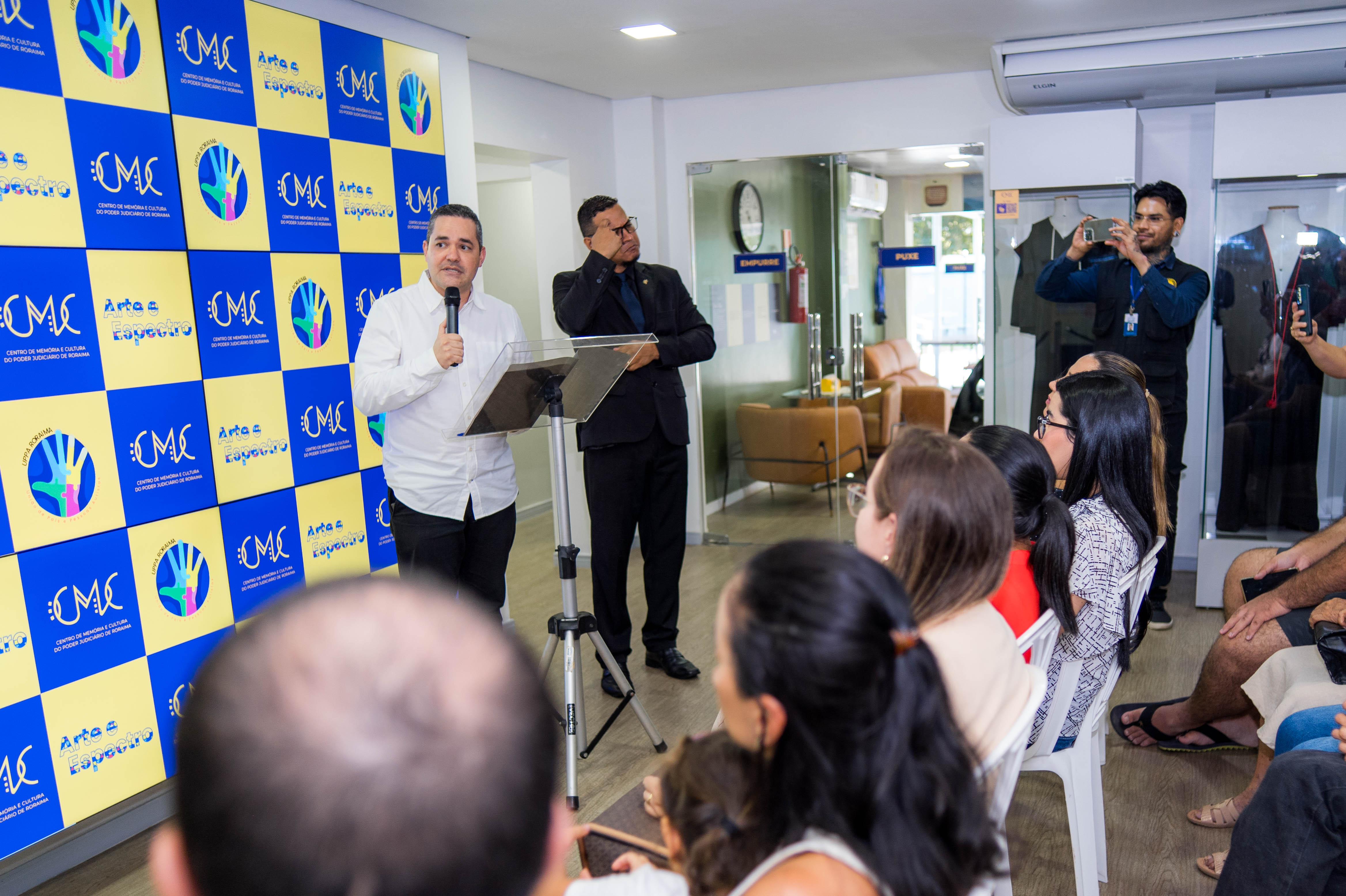 A imagem captura o evento de inicio da exposição. No centro da imagem um homem de camisa branca e calças pretas está falando ao microfone em um púlpito com um tripé. A frente dele algumas pessoas sentadas assistem. Ao seu lado um interprete de libras.