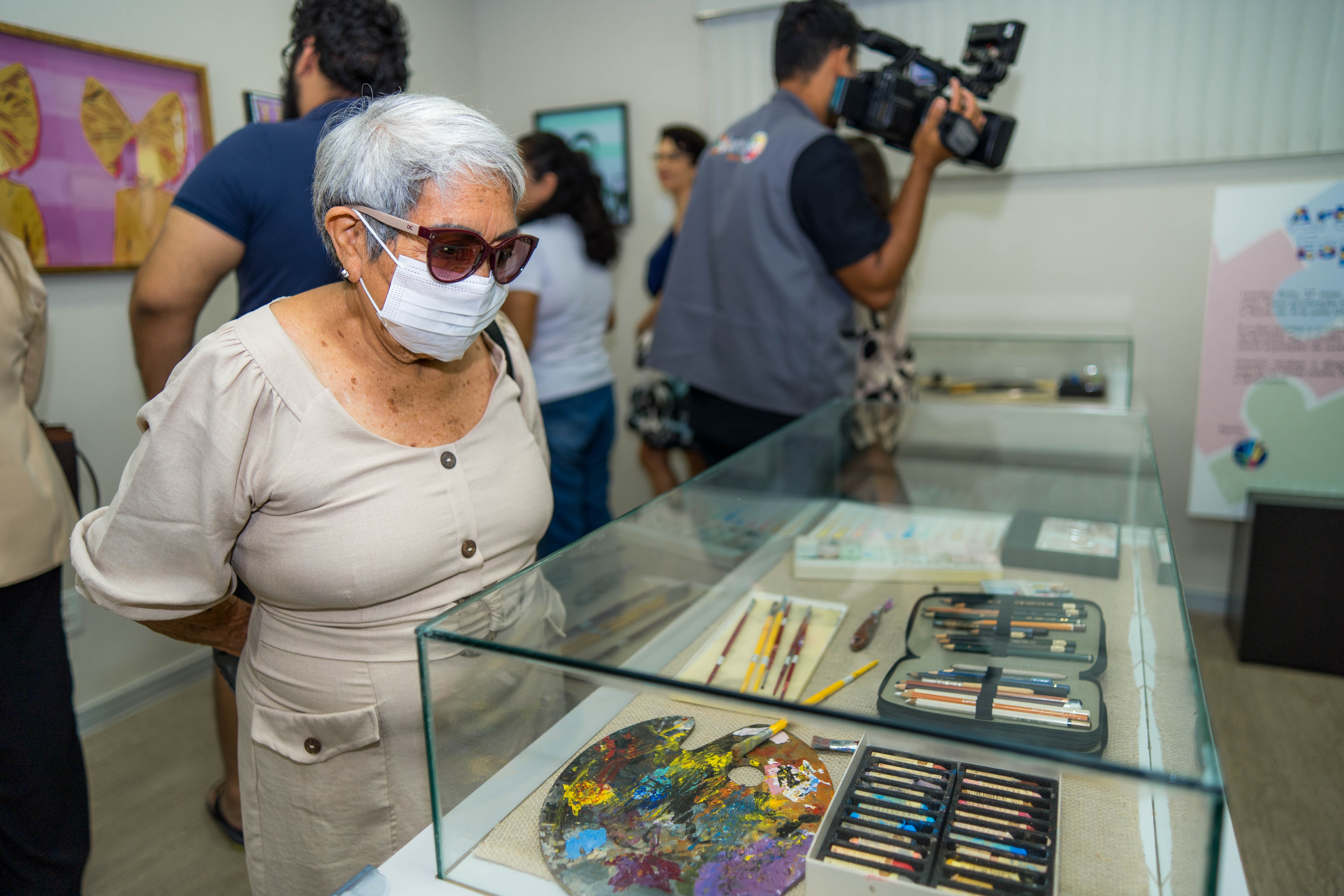 A imagem mostra o interior de uma sala de exposição, onde um grupo de pessoas observa itens em uma vitrine de vidro. Uma senhora com cabelos brancos curtos, óculos escuros e uma máscara facial branca está em primeiro plano, olhando atentamente para o conteúdo da vitrine. Dentro da vitrine, há diversos materiais de arte, incluindo uma paleta de pintura colorida com tinta seca, pincéis de diferentes tamanhos, estojos com lápis de cor e outros materiais de desenho. 