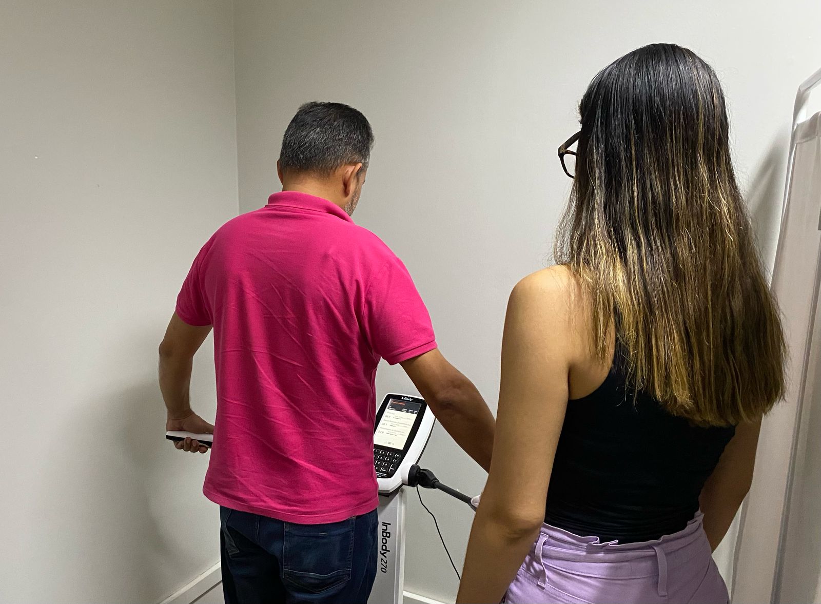 Imagem colorida mostrando a realização d euma avaliação de bioimpedância. Na imagem aparecem duas pessoas. Um homem de costas para a câmera, usando uma camisa polo rosa e calças jeans azuis. Ele está segurando um dispositivo portátil da máquina InBody em sua mão esquerda, com a mão direita na lateral do corpo. Seu cabelo é curto e grisalho. Uma mulher de costas para a câmera, usando um top preto e calças lilás. Seu cabelo é longo e castanho com mechas mais claras, e ela usa óculos pretos. A parede ao fundo é pintada de um tom claro, possivelmente branco ou bege. Uma porta branca pode ser vista à direita da imagem, e parte de outra parede é visível à esquerda. Imagem colorida mostrando a realização d euma avaliação de bioimpedância. Na imagem aparecem duas pessoas. Um homem de costas para a câmera, usando uma camisa polo rosa e calças jeans azuis. Ele está segurando um dispositivo portátil da máquina InBody em sua mão esquerda, com a mão direita na lateral do corpo. Seu cabelo é curto e grisalho. Uma mulher de costas para a câmera, usando um top preto e calças lilás. Seu cabelo é longo e castanho com mechas mais claras, e ela usa óculos pretos. A parede ao fundo é pintada de um tom claro, possivelmente branco ou bege. Uma porta branca pode ser vista à direita da imagem, e parte de outra parede é visível à esquerda.