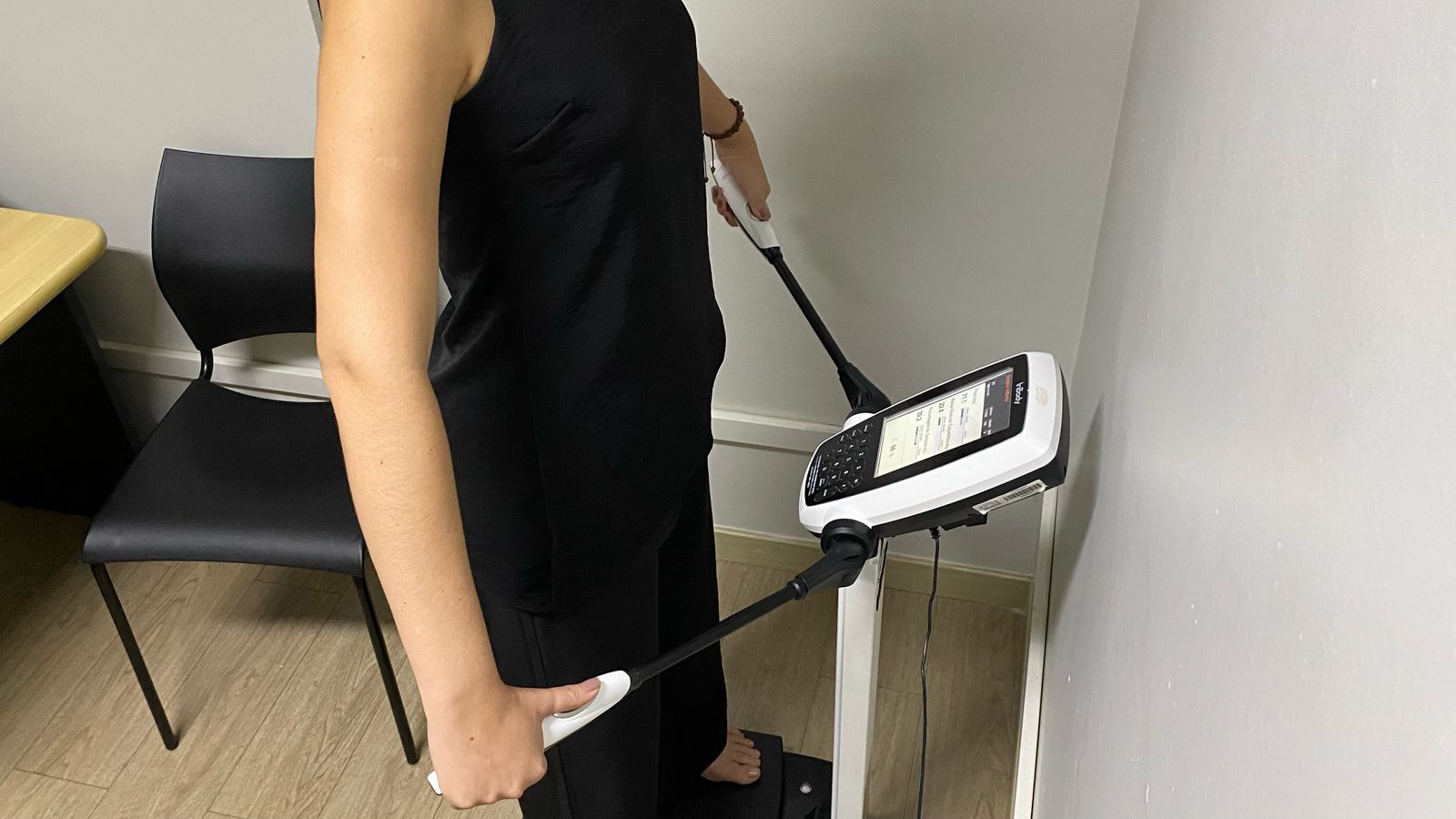 A imagem mostra uma mulher de pé, de lado, em frente a uma máquina de bioimpedância InBody 270. A mulher usa um vestido preto sem mangas e está descalça sobre a plataforma da máquina. Ela está segurando os eletrodos da máquina com ambas as mãos. Uma pulseira dourada é visível em seu pulso direito. A imagem mostra uma mulher de pé, de lado, em frente a uma máquina de bioimpedância InBody 270. A mulher usa um vestido preto sem mangas e está descalça sobre a plataforma da máquina. Ela está segurando os eletrodos da máquina com ambas as mãos. Uma pulseira dourada é visível em seu pulso direito.