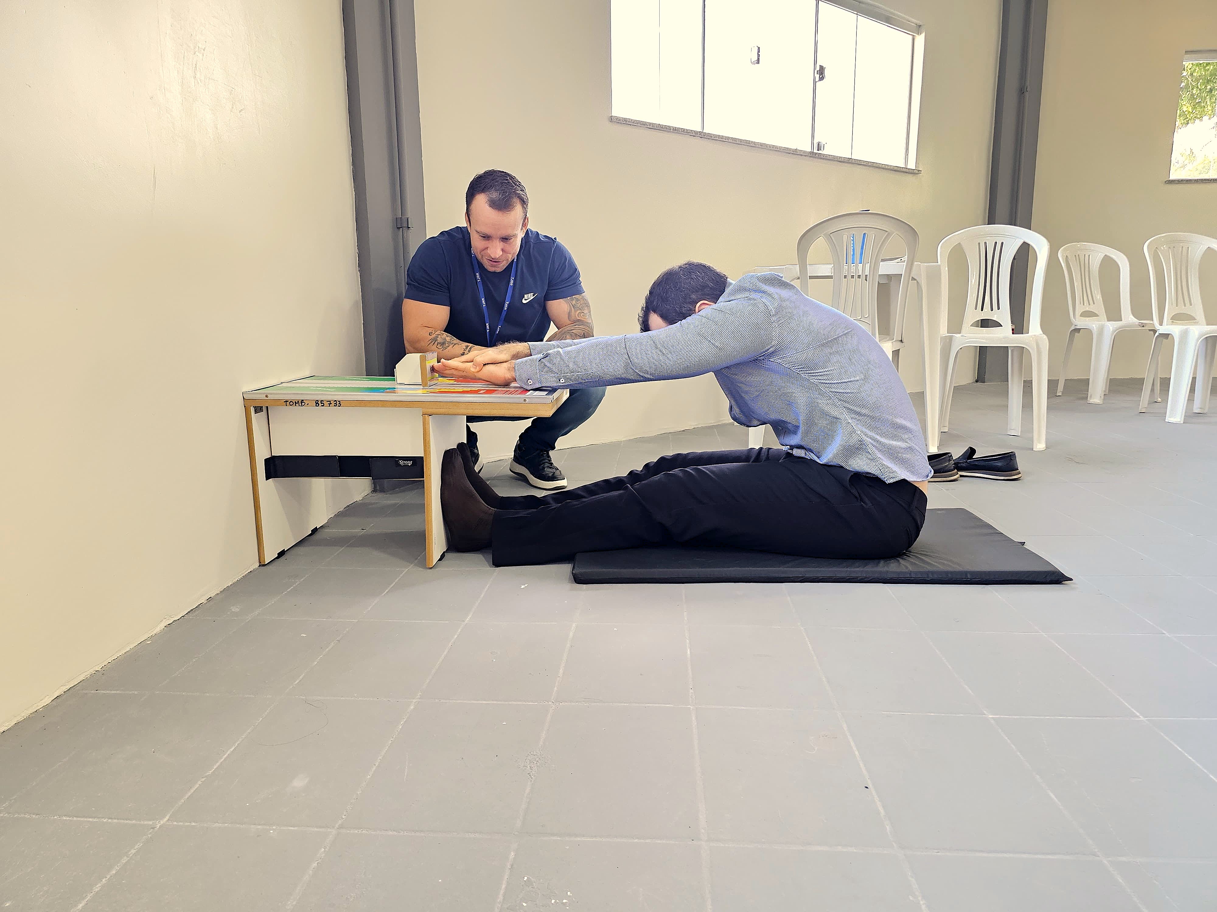 A imagem colorida contém dois homens, sendo um servidor do TJRR fazendo alongamento das pernas com apoio de uma mesinha e do lado dele encontra-se um instrutor de educação física, para ajudar na avaliação e exercício do servidor. Atrás deles encontra-se quatro cadeiras de plástico e uma mesa. 