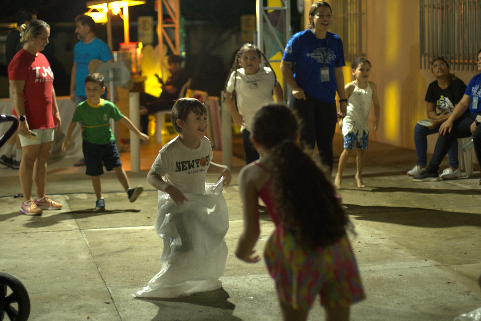 Imagem colorida contém onze pessoas, entre elas cinco crianças brincando de corrida do saco, durante ação alusiva ao Dia Mundial da Infância. Imagem colorida contém onze pessoas, entre elas cinco crianças brincando de corrida do saco, durante ação alusiva ao Dia Mundial da Infância.
