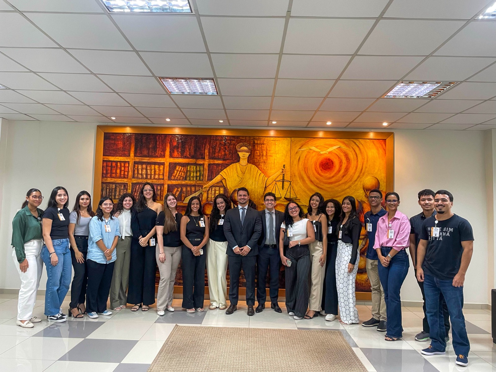 Foto colorida mostra um grupo de dezenove pessoas posando para uma foto em um ambiente interno, provavelmente um prédio público ou uma instituição, dada a presença de uma grande obra de arte na parede ao fundo. Foto colorida mostra um grupo de dezenove pessoas posando para uma foto em um ambiente interno, provavelmente um prédio público ou uma instituição, dada a presença de uma grande obra de arte na parede ao fundo.