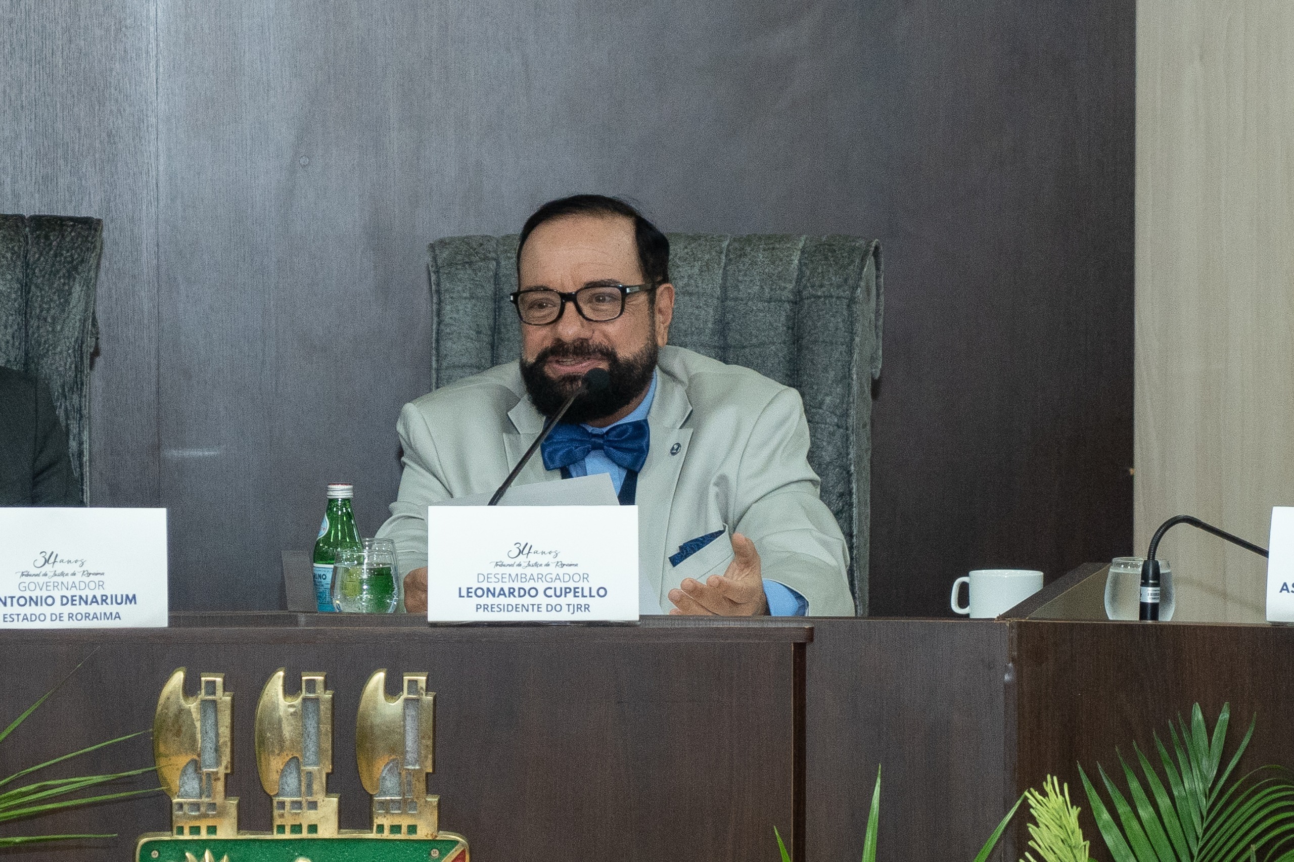 foto colorida do Presidente do TJRR, Leonardo Cupello, sentado atrás de uma mesa com um microfone. Na sua frente há um cartão de papel na mesa que diz "LEONARDO CUPPELLO PRESIDENTE DO TJRR.”