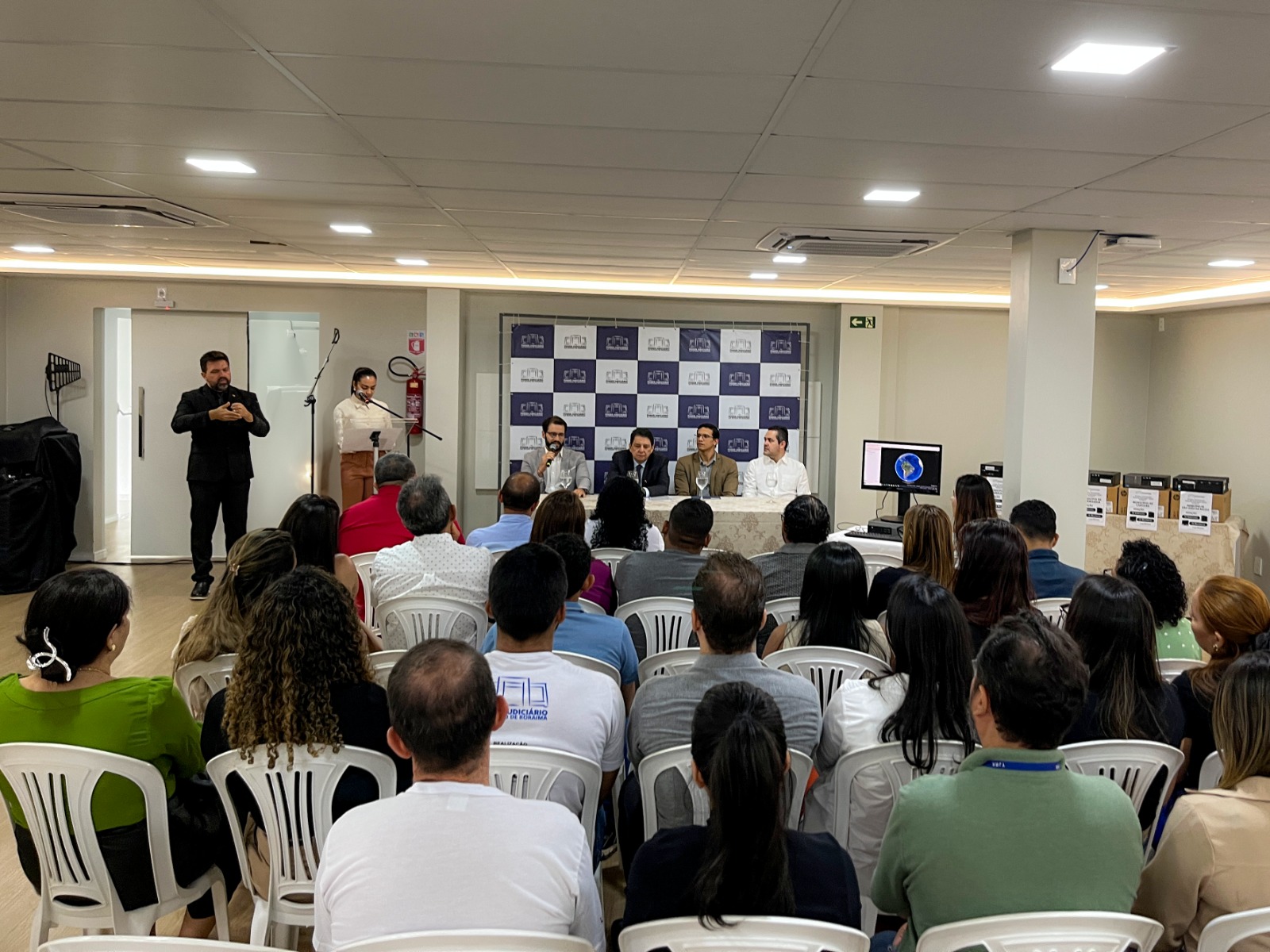 Foto colorida mostra um evento formal de doação de computadores. No centro, há uma mesa com cinco pessoas sentadas, aparentando ser autoridades ou organizadores, em frente a um painel de fundo personalizado com logotipos. À frente deles, um público de aproximadamente 30  pessoas está sentado em cadeiras brancas de plástico, atento à apresentação. À esquerda, um homem de preto faz interpretação em Libras (Língua Brasileira de Sinais), enquanto uma mulher fala ao microfone em um púlpito.