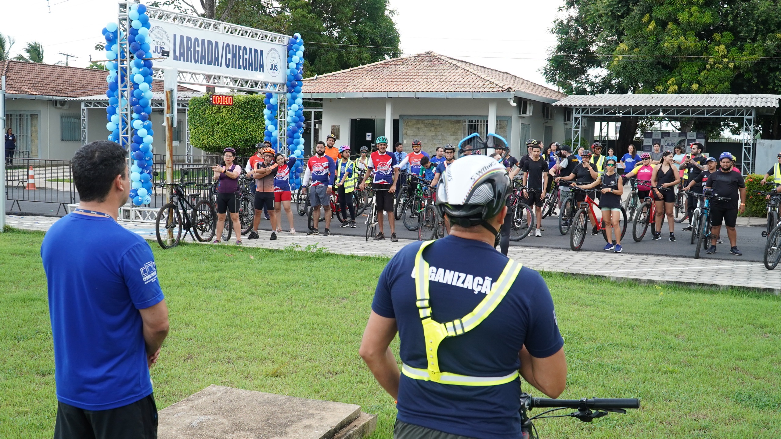 Imagem colorida mostra dezenas de ciclistas se aquecendo em frente à sede da Secretaria de Qualidade de Vida, localizada no Conjunto Sociocultural do TJRR. Ao lado dos ciclistas, há uma placa indicando o local de largada e chegada.