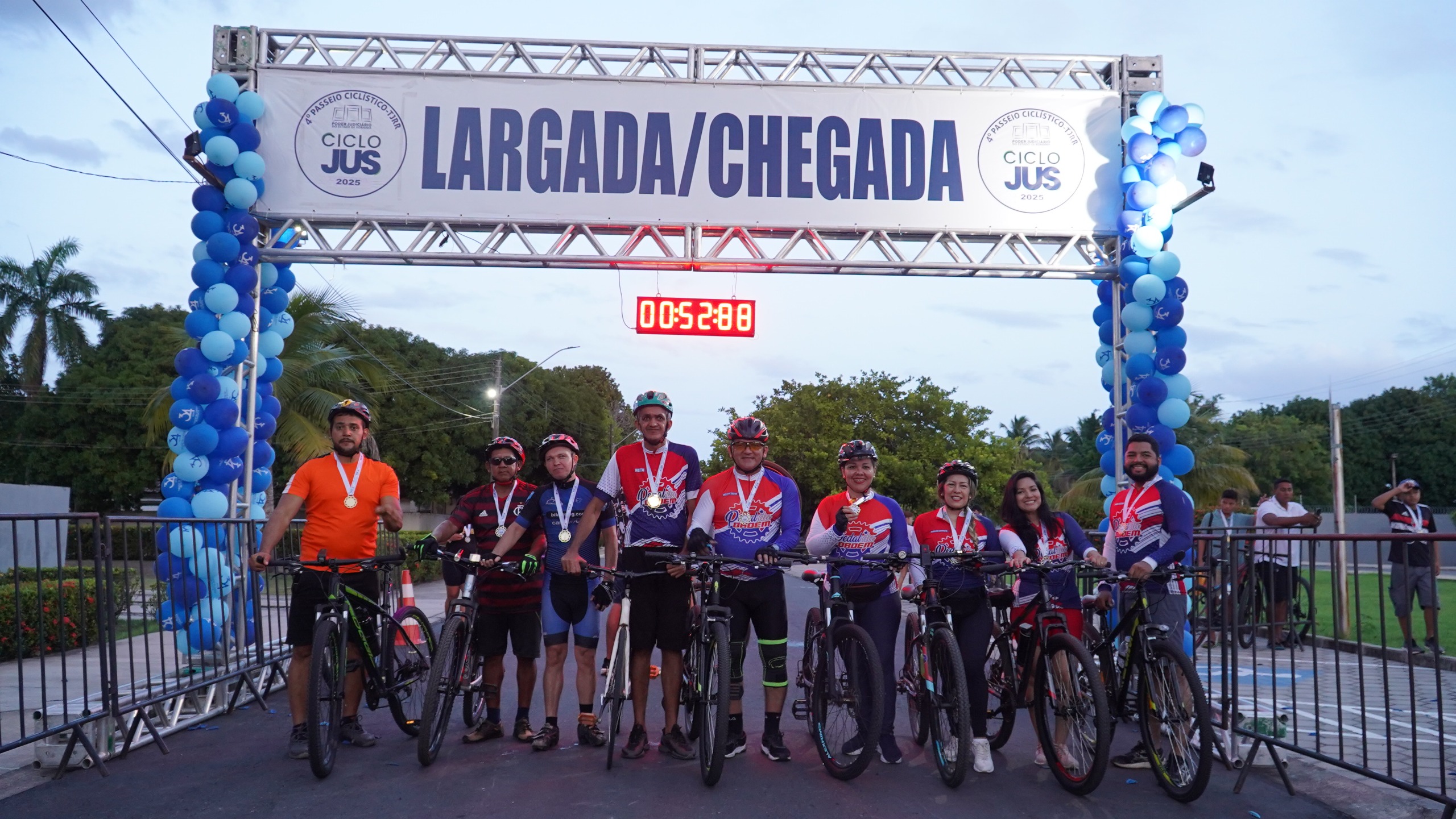 Imagem colorida mostra o grupo de ciclistas "Pedal da Ordem", com nove integrantes posando para foto no ponto de largada e chegada do IV CicloJus do TJRR.