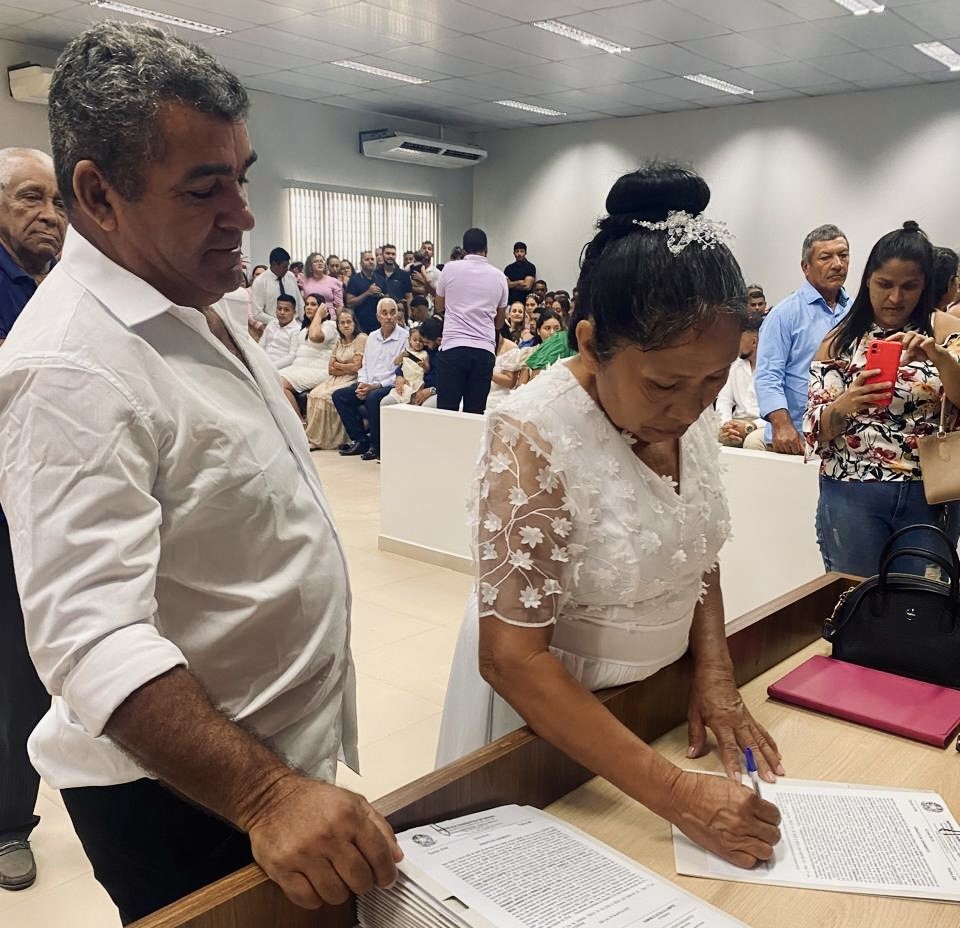Foto colorida de uma noiva, vestida de branco ao lado do seu esposo assinando um papel.