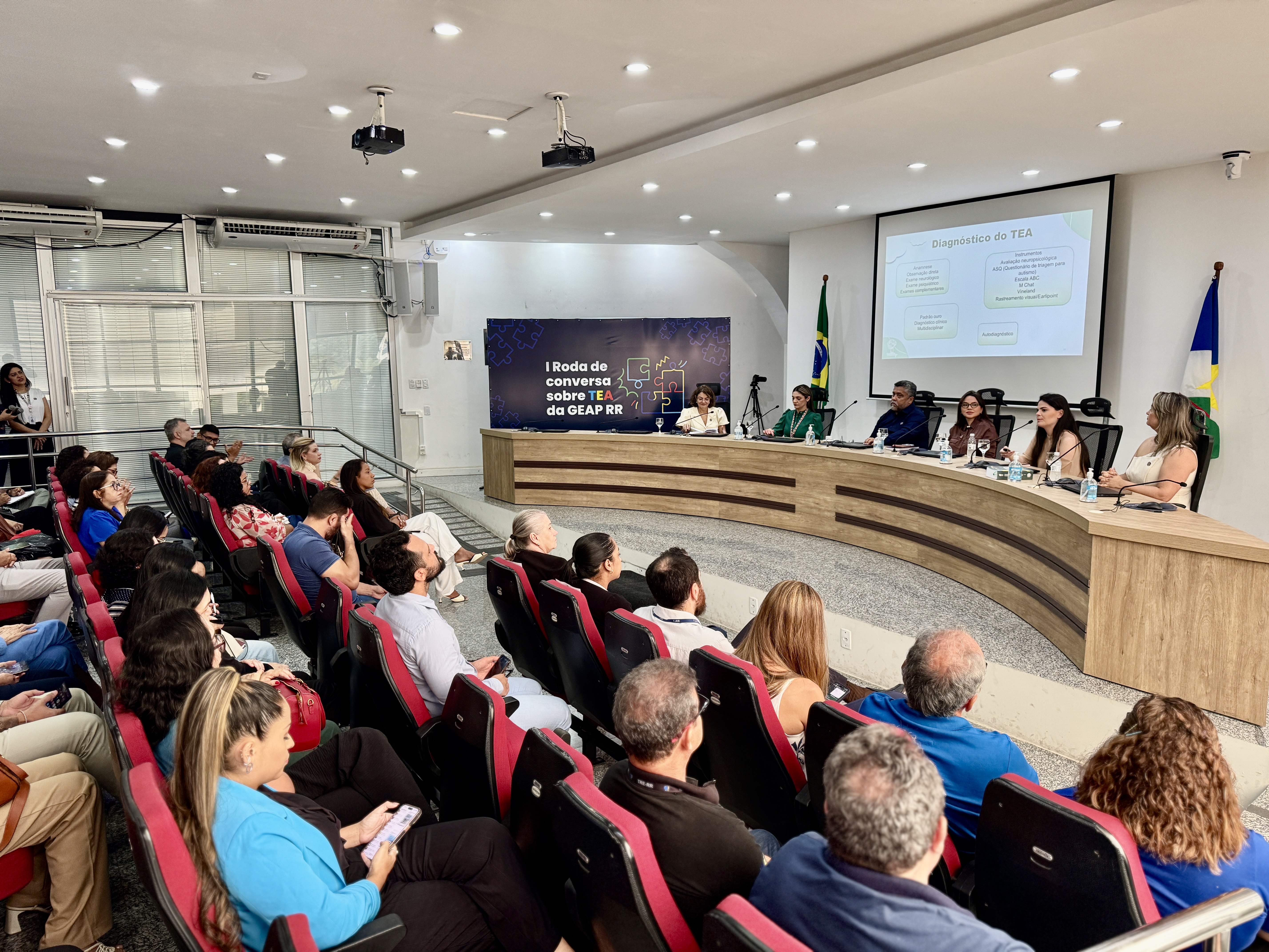 Foto colorida contém publico sentado assistindo palestrantes que abordaram o TEA em Roraima, no auditório do Tribunal Regional Eleitoral (TRE-RR). Os palestrantes se encontram sentados em uma mesa de frente pra plateia, e atrás deles está um slide com o titulo “Diagnóstico do TEA”. Foto colorida contém publico sentado assistindo palestrantes que abordaram o TEA em Roraima, no auditório do Tribunal Regional Eleitoral (TRE-RR). Os palestrantes se encontram sentados em uma mesa de frente pra plateia, e atrás deles está um slide com o titulo “Diagnóstico do TEA”.