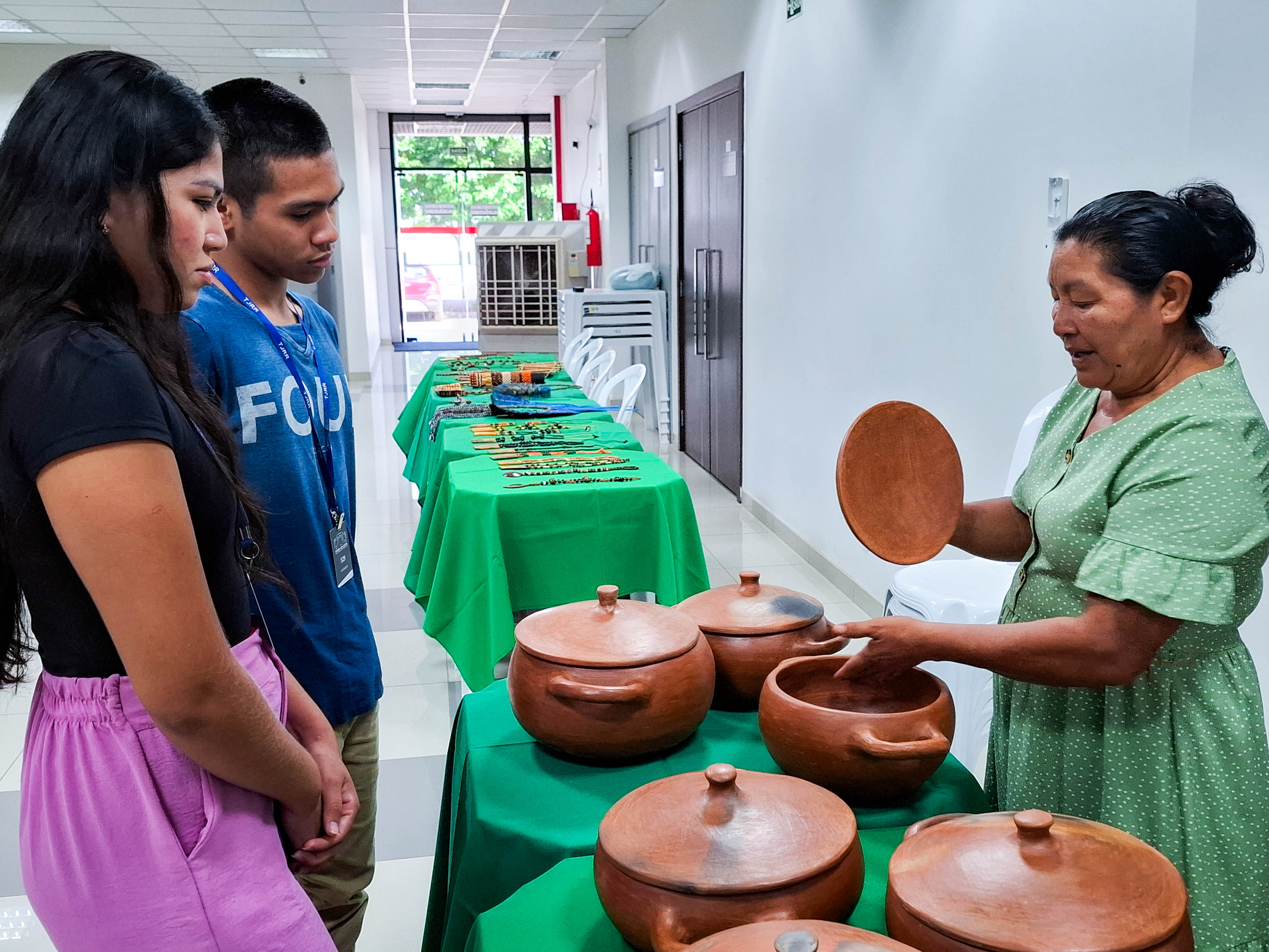 A imagem colorida mostra três indivíduos reunidos em torno de uma mesa coberta por uma toalha verde vibrante. Uma jovem está sentada à esquerda, enquanto um homem e uma mulher estão em pé à direita da mesa. 