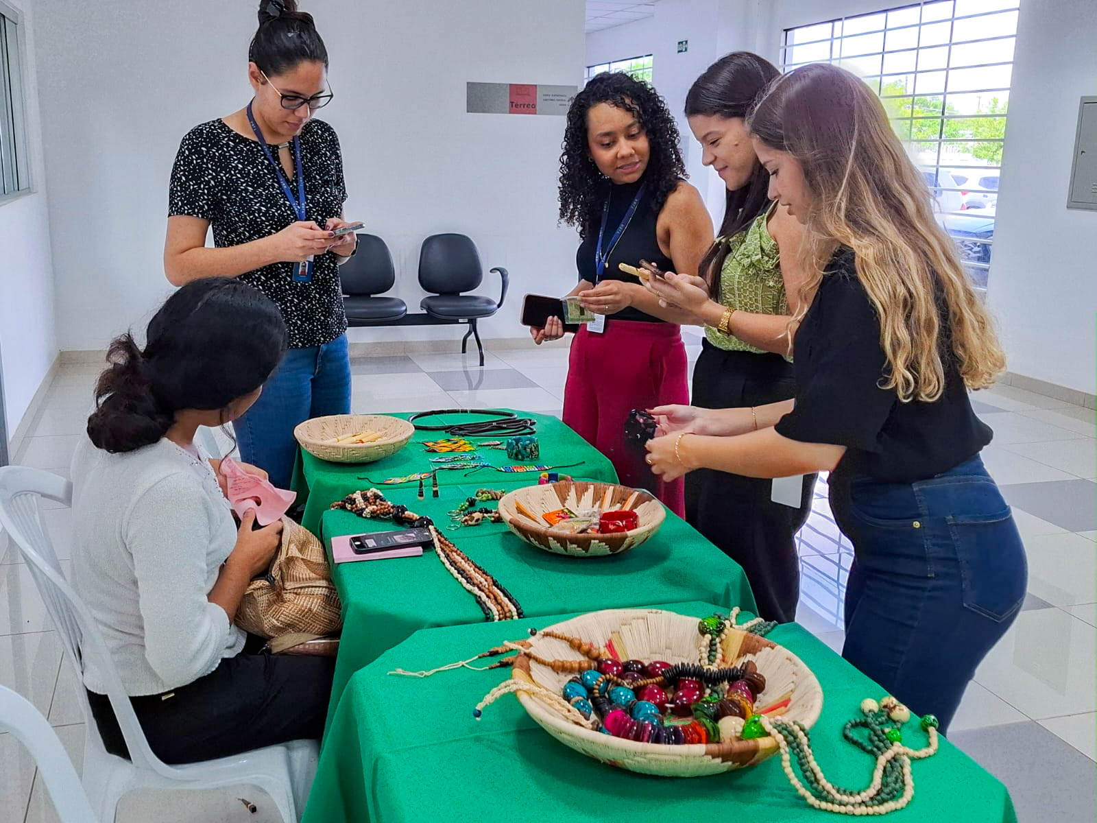 A imagem colorida mostra cinco mulheres reunidas em torno de uma mesa coberta por uma toalha de cor verde vibrante. Quatro mulheres estão em pé, observando e interagindo com os objetos dispostos sobre a mesa, enquanto uma quinta mulher está sentada à esquerda, de costas para a câmera, trabalhando em algo com as mãos. Na mesa, há diversos itens que parecem ser materiais para artesanato indígena.