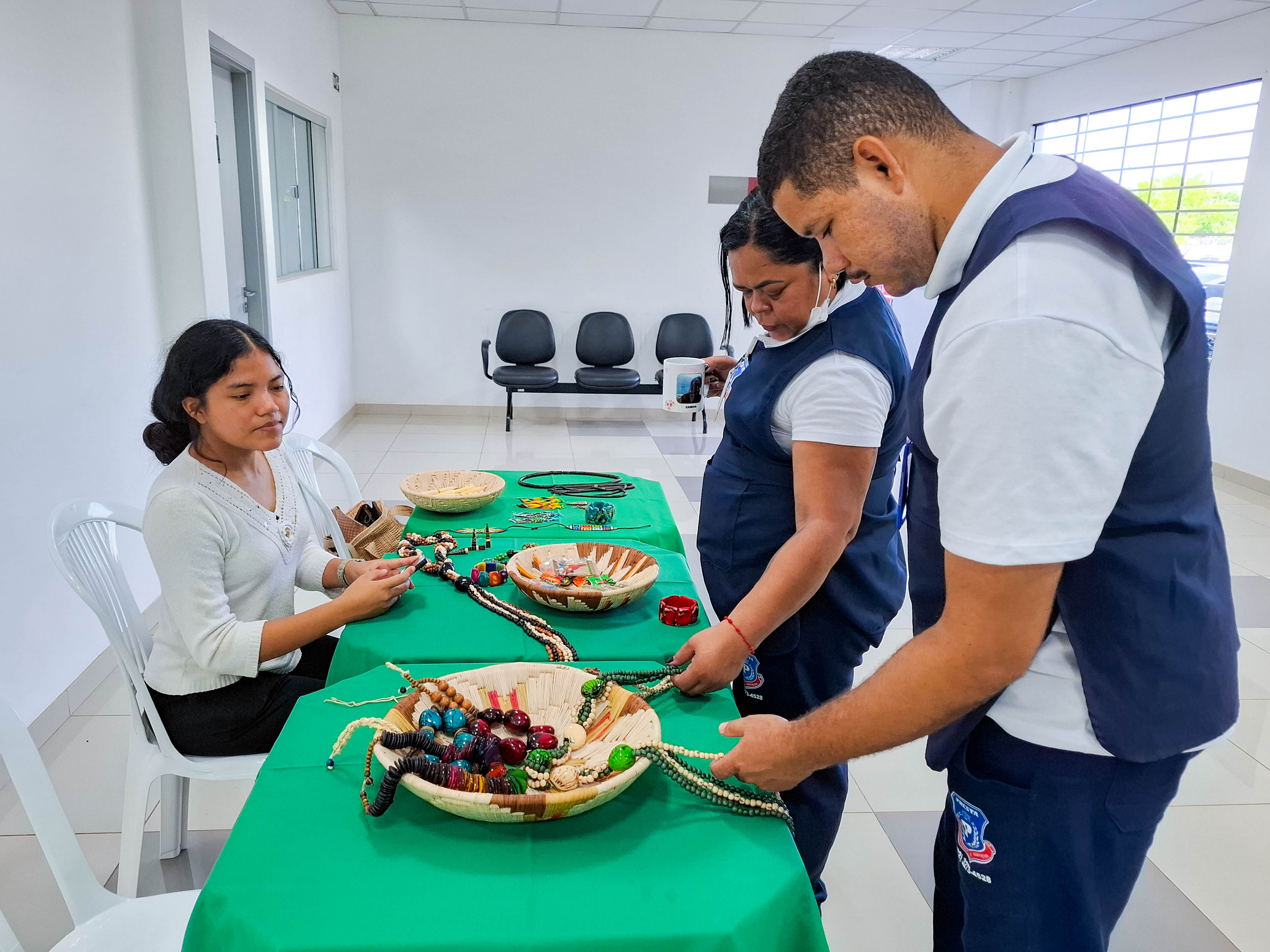  imagem colorida mostra três pessoas reunidas em torno de uma mesa coberta por uma toalha verde. Uma jovem está sentada à esquerda, enquanto um homem e uma mulher estão em pé à direita da mesa.