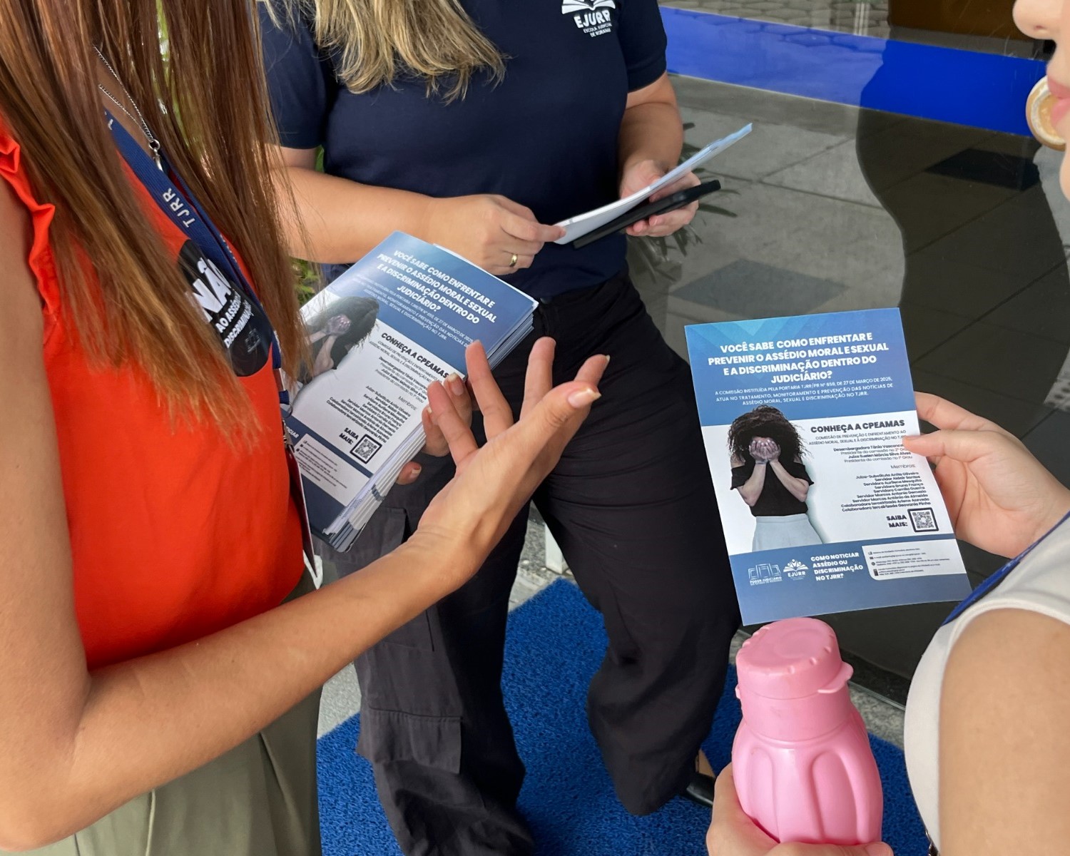 Foto colorida mostra um momento de conversa entre três mulheres em frente a um prédio, sobre um tapete azul. O foco está no panfleto que uma delas segura, um material informativo sobre como enfrentar, prevenir e denunciar o assédio moral, sexual e discriminação no ambiente do Judiciário. Foto colorida mostra um momento de conversa entre três mulheres em frente a um prédio, sobre um tapete azul. O foco está no panfleto que uma delas segura, um material informativo sobre como enfrentar, prevenir e denunciar o assédio moral, sexual e discriminação no ambiente do Judiciário.