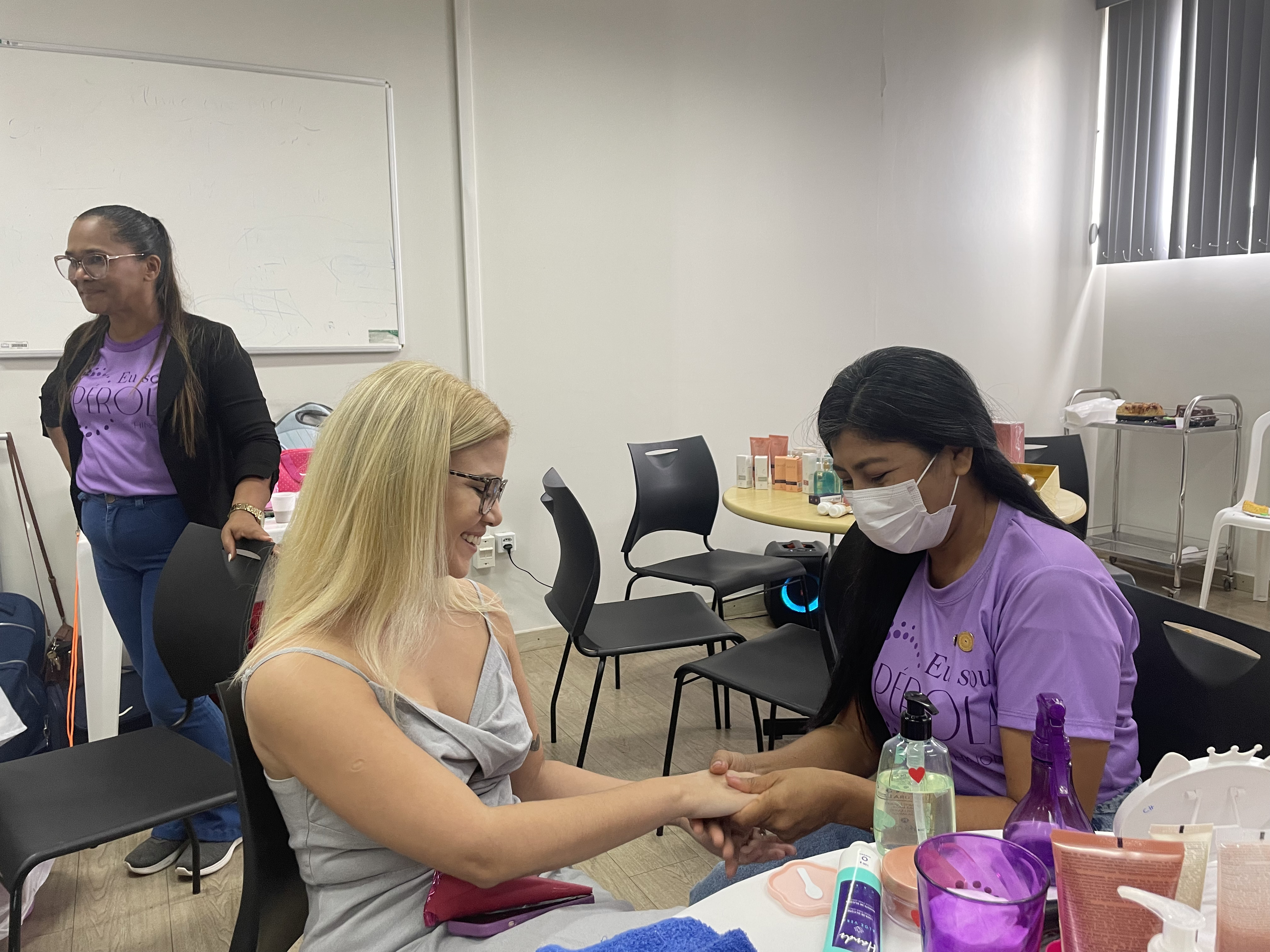 Imagem colorida de uma servidora sorrindo enquanto faz o spa das mãos com uma mulher da equipe de beleza na Blitz da Beleza. Imagem colorida de uma servidora sorrindo enquanto faz o spa das mãos com uma mulher da equipe de beleza na Blitz da Beleza.
