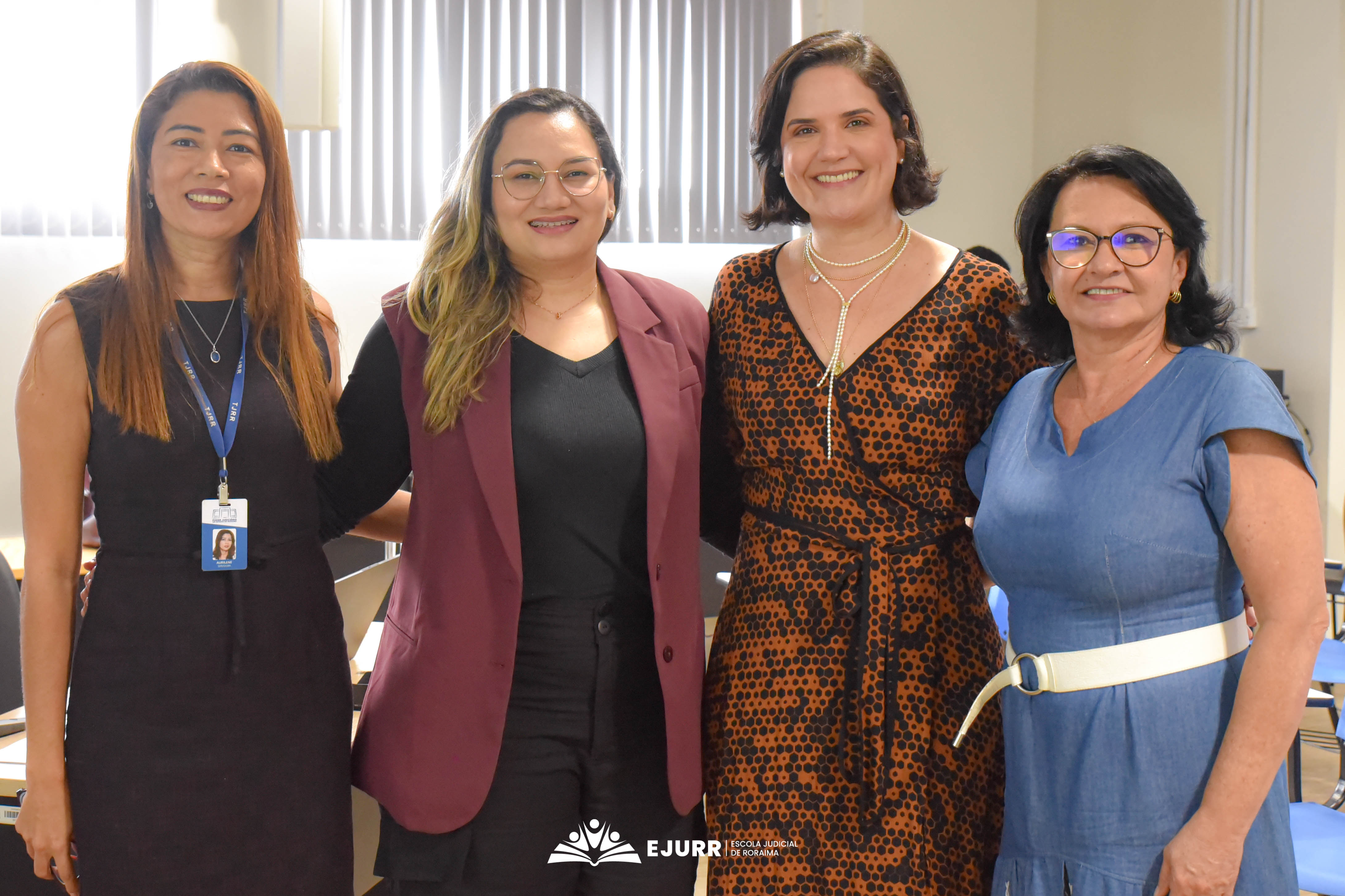 A imagem apresenta quatro mulheres, que sorriem e estão abraçadas para a foto. A primeira usa crachá, vestido preto e tem cabelos compridos; a segunda tem cabelos compridos, usa óculos e veste roupa preta e um colete na cor vinho; a terceira mulher, é a palestrante, que possui cabelos médios e escuros, usa colar e vestido; a quarta possui cabelos médios escuros, usa óculos, vestido azul e cinto branco. Na parte inferior, está a logo da Ejurr com letras brancas. A imagem apresenta quatro mulheres, que sorriem e estão abraçadas para a foto. A primeira usa crachá, vestido preto e tem cabelos compridos; a segunda tem cabelos compridos, usa óculos e veste roupa preta e um colete na cor vinho; a terceira mulher, é a palestrante, que possui cabelos médios e escuros, usa colar e vestido; a quarta possui cabelos médios escuros, usa óculos, vestido azul e cinto branco. Na parte inferior, está a logo da Ejurr com letras brancas.