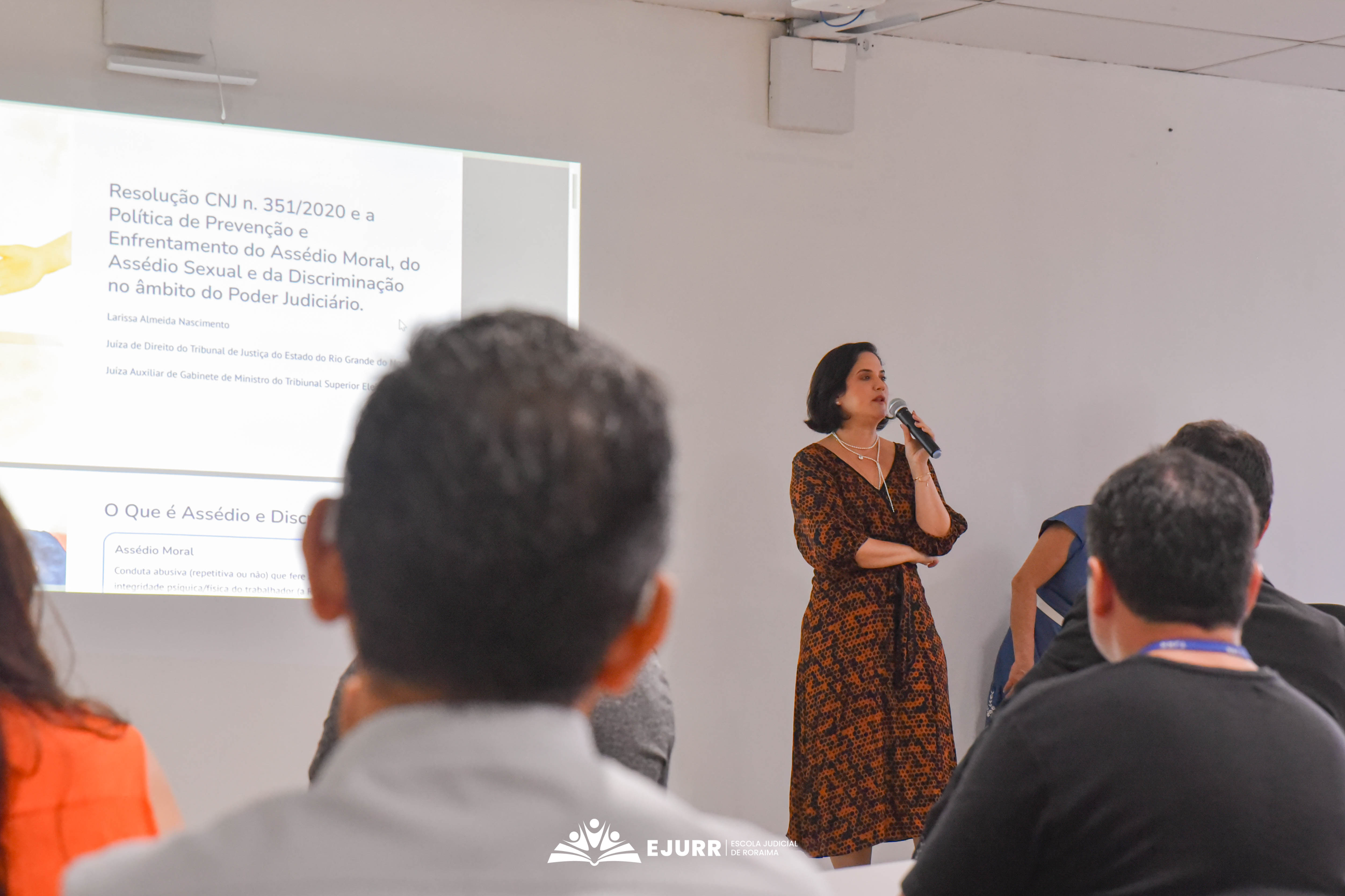 A imagem apresenta a palestrante que utiliza microfone, tendo ao fundo o título da palestra projetada em uma parede branca. Ela usa vestido em tons marrom e laranja. No primeiro plano, estão pessoas sentadas de costas. Na parte inferior está a logo da Ejurr na cor branca. A imagem apresenta a palestrante que utiliza microfone, tendo ao fundo o título da palestra projetada em uma parede branca. Ela usa vestido em tons marrom e laranja. No primeiro plano, estão pessoas sentadas de costas. Na parte inferior está a logo da Ejurr na cor branca.