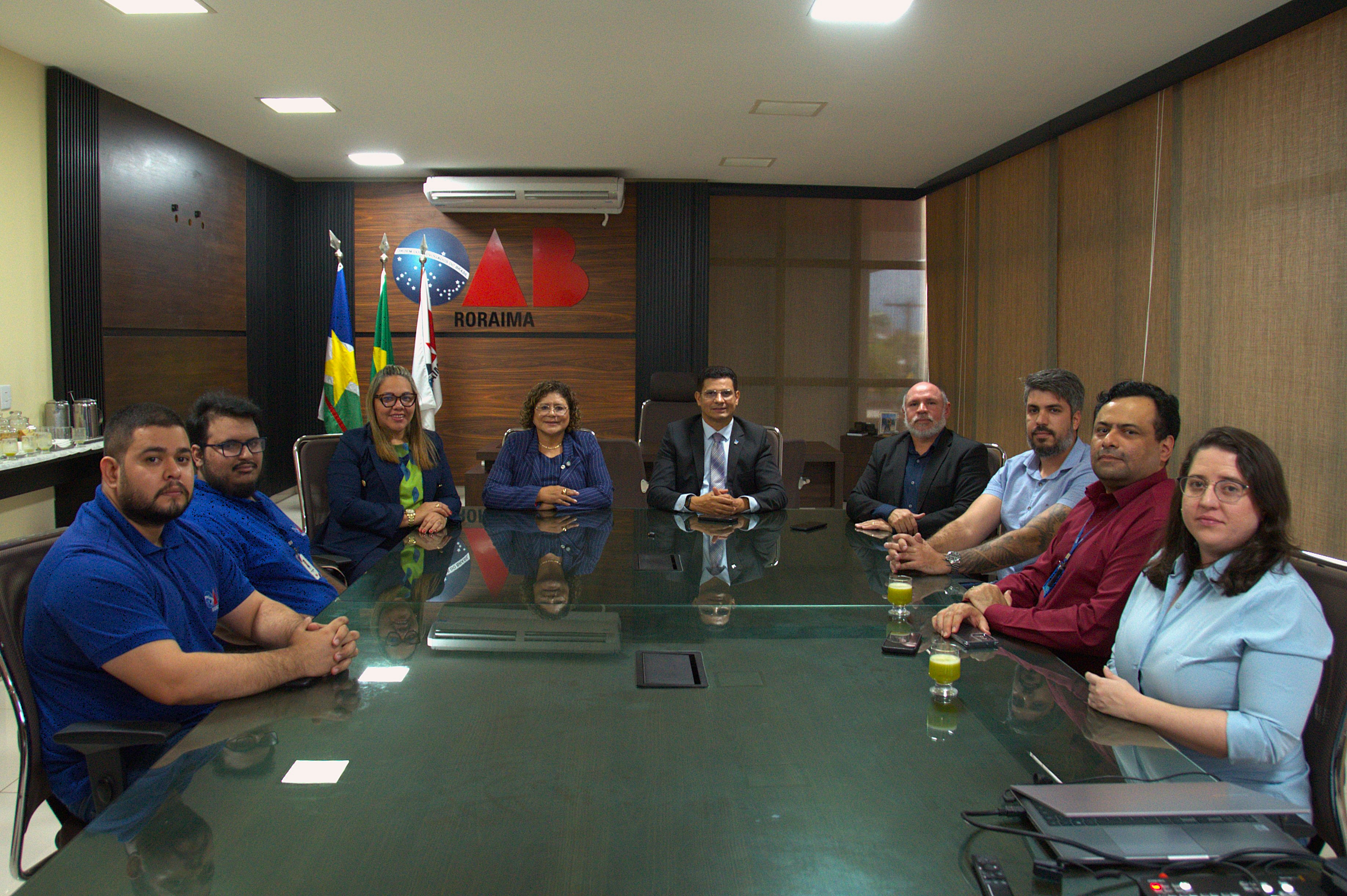 A imagem mostra oito pessoas reunidas ao redor de uma mesa de conferência de vidro, posando para foto em um ambiente formal. Ao fundo, estão as bandeiras do Brasil, de Roraima e uma terceira não identificada, além do logotipo "Roráima". A cena sugere um encontro institucional ou governamental. A imagem mostra oito pessoas reunidas ao redor de uma mesa de conferência de vidro, posando para foto em um ambiente formal. Ao fundo, estão as bandeiras do Brasil, de Roraima e uma terceira não identificada, além do logotipo "Roráima". A cena sugere um encontro institucional ou governamental.