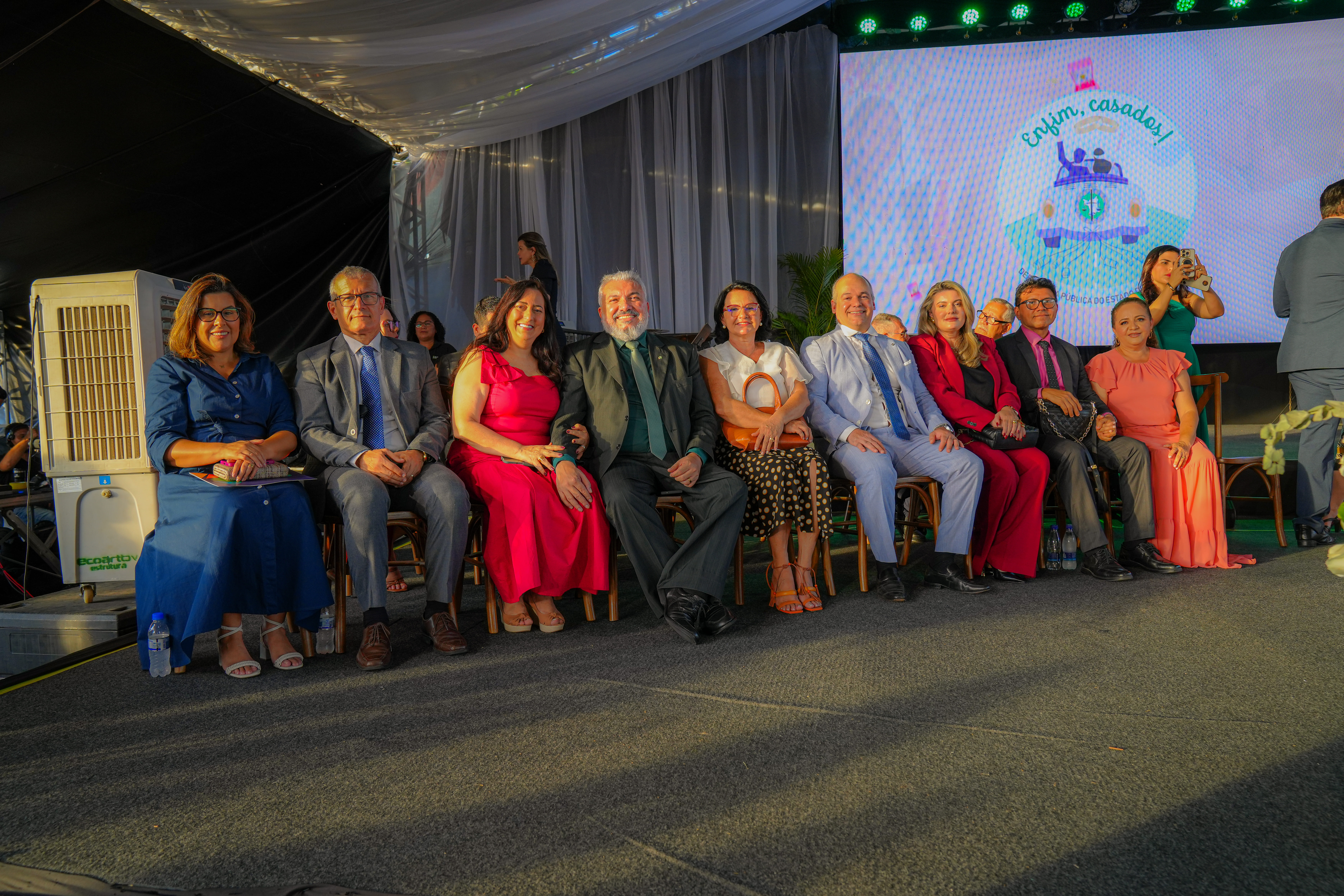 Foto colorida mostra um grupo de pessoas sentadas em uma fila, sob uma tenda, com um telão ao fundo. Há 5 mulheres e 4 homens de várias idades, todos vestidos com trajes de gala. Entre eles, o Defensor Público-Geral, Oleno Matos; a Diretora da EJURR, Desembargadora Tânia Vasconcelos; o Juiz corregedor do TJRR, Eduardo Carvalho e a Defensora pública, Elceni Diogo. No telão, pode-se ver a frase "Enfim, casados!" e uma imagem de um carro de casamento. Foto colorida mostra um grupo de pessoas sentadas em uma fila, sob uma tenda, com um telão ao fundo. Há 5 mulheres e 4 homens de várias idades, todos vestidos com trajes de gala. Entre eles, o Defensor Público-Geral, Oleno Matos; a Diretora da EJURR, Desembargadora Tânia Vasconcelos; o Juiz corregedor do TJRR, Eduardo Carvalho e a Defensora pública, Elceni Diogo. No telão, pode-se ver a frase "Enfim, casados!" e uma imagem de um carro de casamento.