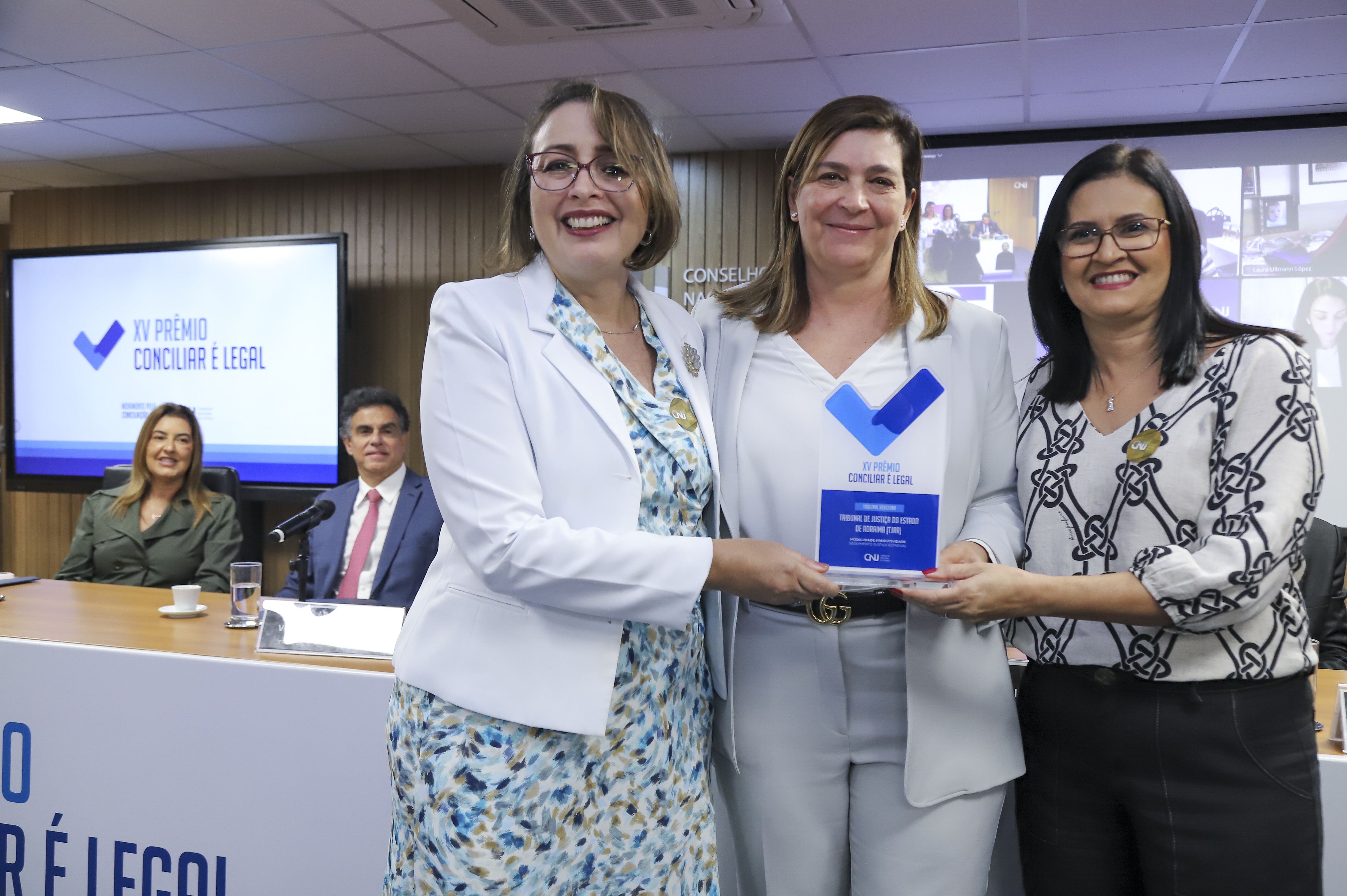A imagem registra um momento de premiação durante o XV Prêmio Conciliar é Legal. Três mulheres sorriem em primeiro plano, com duas delas segurando um troféu azul e branco. O cenário é um auditório, com painel de madeira, mesa oficial ao fundo e projeção do nome do prêmio em destaque. À esquerda está a juíza auxiliar da Presidência, Lana Leitão, e à direita, a servidora Ocimara Vasconcelos. A imagem registra um momento de premiação durante o XV Prêmio Conciliar é Legal. Três mulheres sorriem em primeiro plano, com duas delas segurando um troféu azul e branco. O cenário é um auditório, com painel de madeira, mesa oficial ao fundo e projeção do nome do prêmio em destaque. À esquerda está a juíza auxiliar da Presidência, Lana Leitão, e à direita, a servidora Ocimara Vasconcelos.