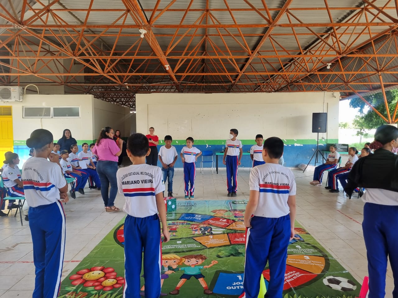 Imagem colorida mostra um grupo de crianças, adolescentes e alguns adultos em um pátio escolar coberto. No centro, há um grande tapete de chão que se assemelha a um jogo de tabuleiro com ilustrações coloridas. Vários estudantes, vestindo uniformes azuis e brancos, estão em pé ao redor do tapete, alguns olhando para a câmera e outros para o jogo. Há também estudantes sentados em cadeiras de plástico ao redor da área principal. Dois adultos estão visíveis no lado esquerdo, e o ambiente é caracterizado por um telhado de treliça metálica e pilares. Imagem colorida mostra um grupo de crianças, adolescentes e alguns adultos em um pátio escolar coberto. No centro, há um grande tapete de chão que se assemelha a um jogo de tabuleiro com ilustrações coloridas. Vários estudantes, vestindo uniformes azuis e brancos, estão em pé ao redor do tapete, alguns olhando para a câmera e outros para o jogo. Há também estudantes sentados em cadeiras de plástico ao redor da área principal. Dois adultos estão visíveis no lado esquerdo, e o ambiente é caracterizado por um telhado de treliça metálica e pilares.