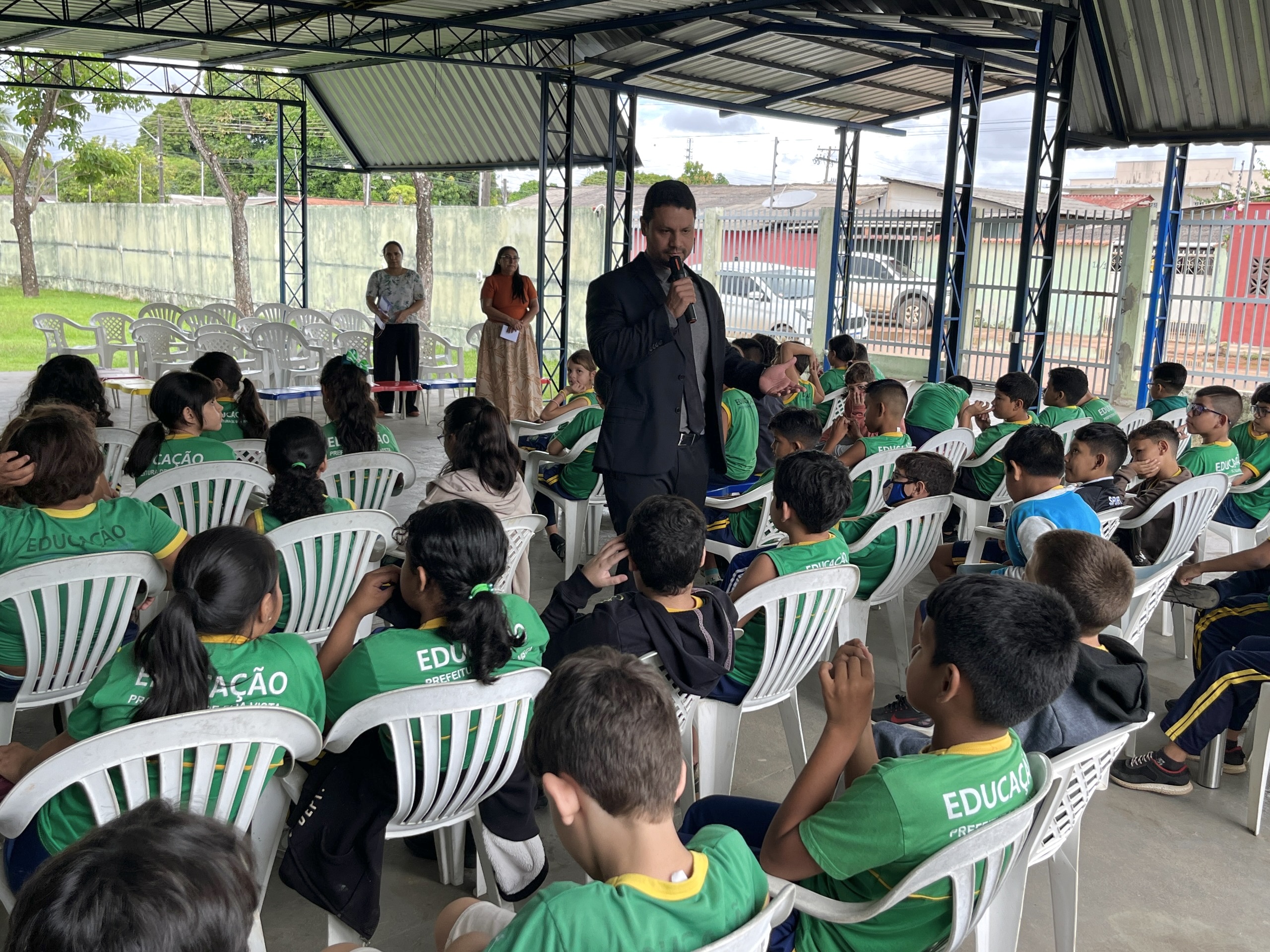 A imagem colorida retrata um evento educacional em um espaço coberto e arejado, com várias crianças vestindo camisetas verdes com a palavra “EDUCAÇÃO”. Elas estão sentadas e atentas, assistindo ao discruso do juiza Cleber Gonçalves, que fala ao microfone. O ambiente é simples, com estrutura metálica e cercado por árvores ao fundo. A imagem colorida retrata um evento educacional em um espaço coberto e arejado, com várias crianças vestindo camisetas verdes com a palavra “EDUCAÇÃO”. Elas estão sentadas e atentas, assistindo ao discruso do juiza Cleber Gonçalves, que fala ao microfone. O ambiente é simples, com estrutura metálica e cercado por árvores ao fundo.