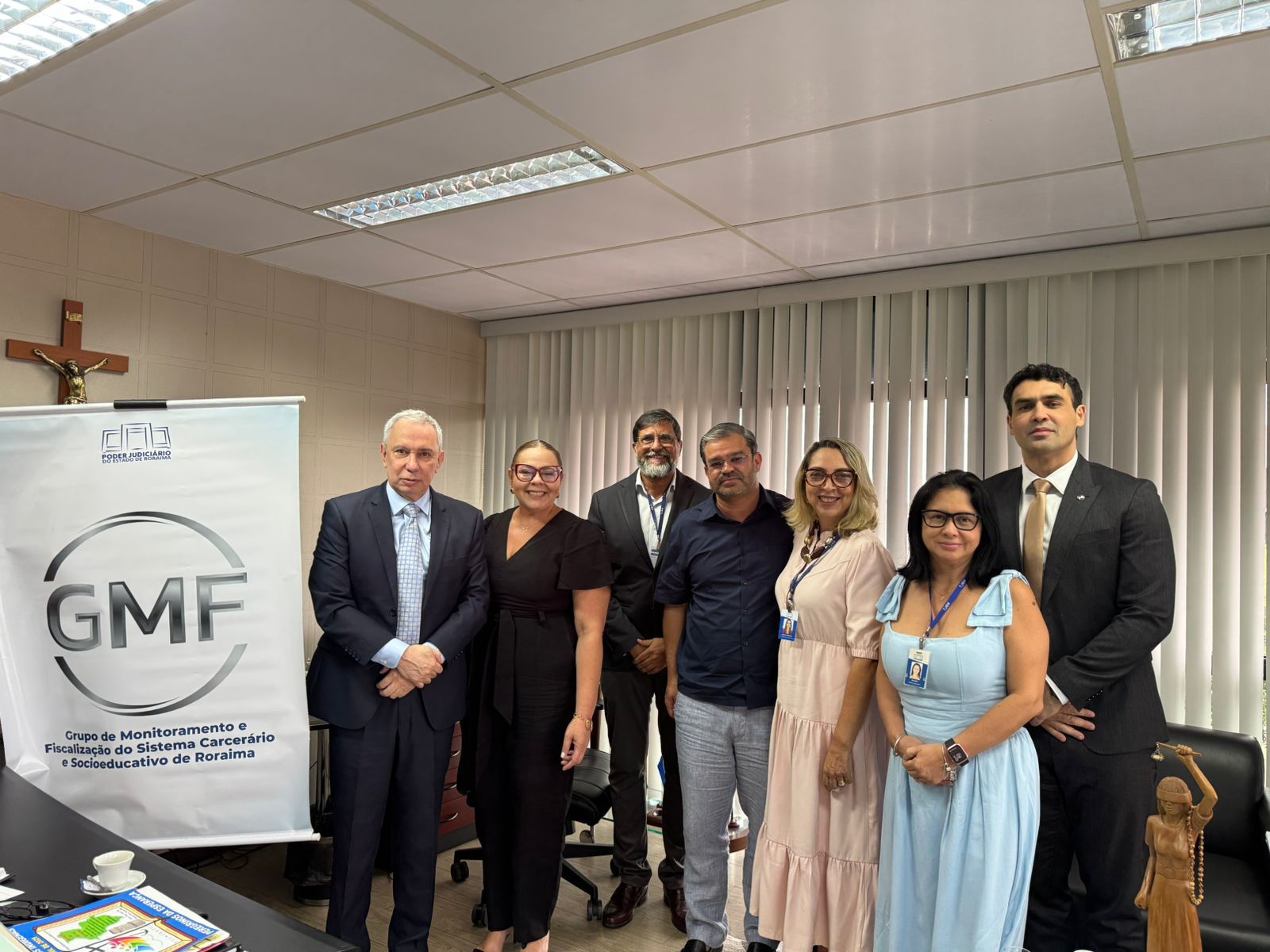 Foto colorida mostra a equipe do GMF, posando pra foto com a equipe da Sesau. A imagem mostra um grupo de sete pessoas, cinco homens e duas mulheres, posando para uma foto em um ambiente interno que parece ser um escritório ou sala de reuniões. À esquerda da imagem, há um banner vertical branco com o logotipo "GMF" em cinza e o texto "Grupo de Monitoramento e Fiscalização do Sistema Carcerário e Socioeducativo de Roraima". Foto colorida mostra a equipe do GMF, posando pra foto com a equipe da Sesau. A imagem mostra um grupo de sete pessoas, cinco homens e duas mulheres, posando para uma foto em um ambiente interno que parece ser um escritório ou sala de reuniões. À esquerda da imagem, há um banner vertical branco com o logotipo "GMF" em cinza e o texto "Grupo de Monitoramento e Fiscalização do Sistema Carcerário e Socioeducativo de Roraima".