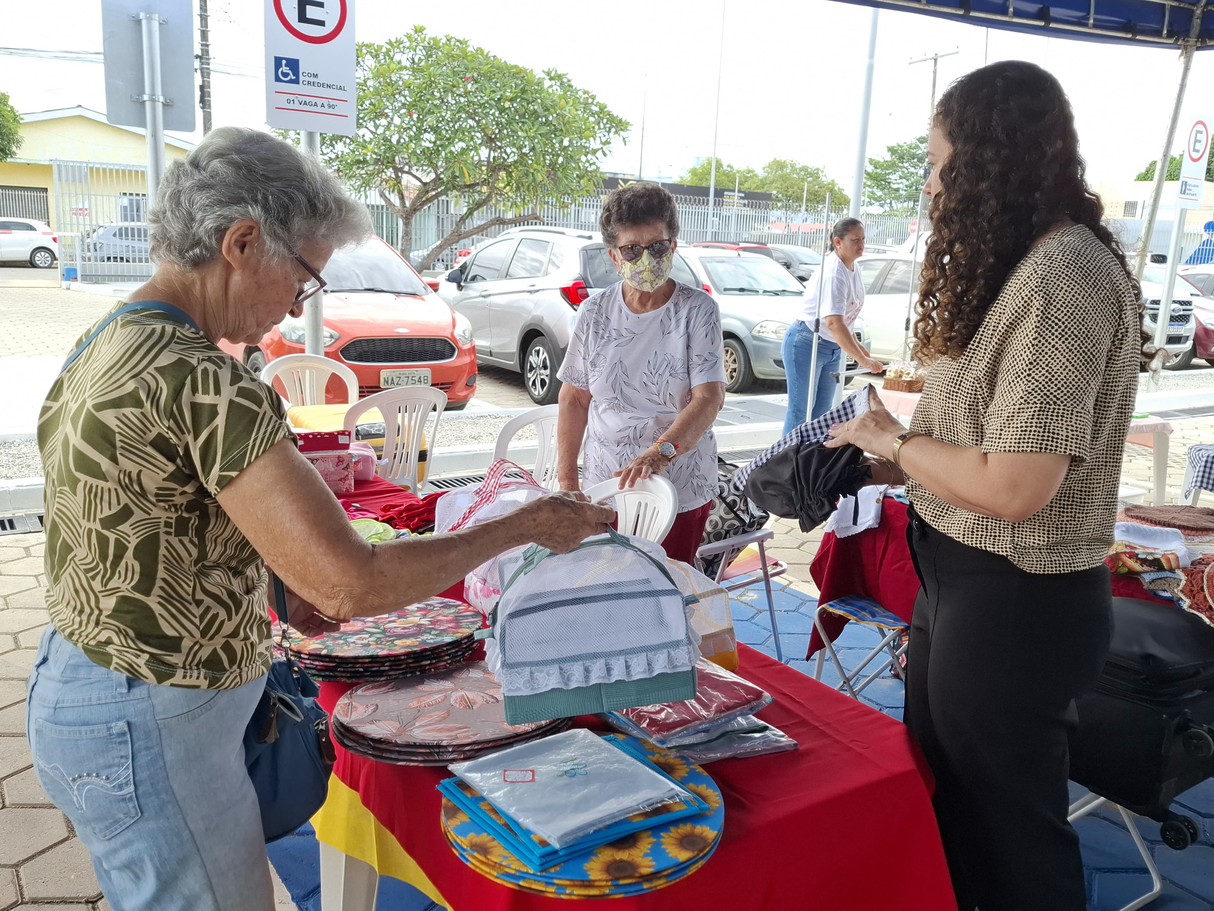 A imagem mostra três mulheres interagindo em uma barraca de feira ao ar livre, em um ambiente ensolarado próximo a um estacionamento. Uma delas, à esquerda, observa os itens expostos, enquanto outra, atrás da barraca, organiza os produtos. À direita, a terceira mulher segura um item, possivelmente de vestuário. A barraca exibe artigos artesanais como jogos americanos coloridos, redes de proteção para alimentos e cestos decorativos. Foto 2: A imagem mostra três mulheres interagindo em uma barraca de feira ao ar livre, em um ambiente ensolarado próximo a um estacionamento. Uma delas, à esquerda, observa os itens expostos, enquanto outra, atrás da barraca, organiza os produtos. À direita, a terceira mulher segura um item, possivelmente de vestuário. A barraca exibe artigos artesanais como jogos americanos coloridos, redes de proteção para alimentos e cestos decorativos. Foto 2: