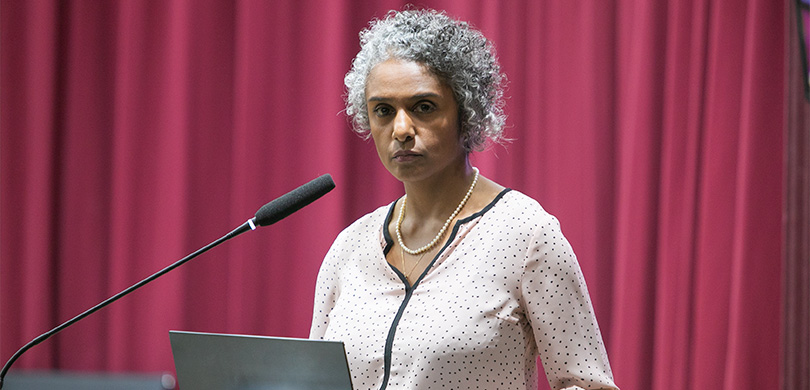 A imagem mostra uma mulher negra, de cabelos cacheados grisalhos. Ela usa cordão dourado e de pérolas, blusa de manga poá com detalhes pretos. Há um microfone à sua frente, um notebook, e atrás, há uma grande cortina na cor vinho. A imagem mostra uma mulher negra, de cabelos cacheados grisalhos. Ela usa cordão dourado e de pérolas, blusa de manga poá com detalhes pretos. Há um microfone à sua frente, um notebook, e atrás, há uma grande cortina na cor vinho.