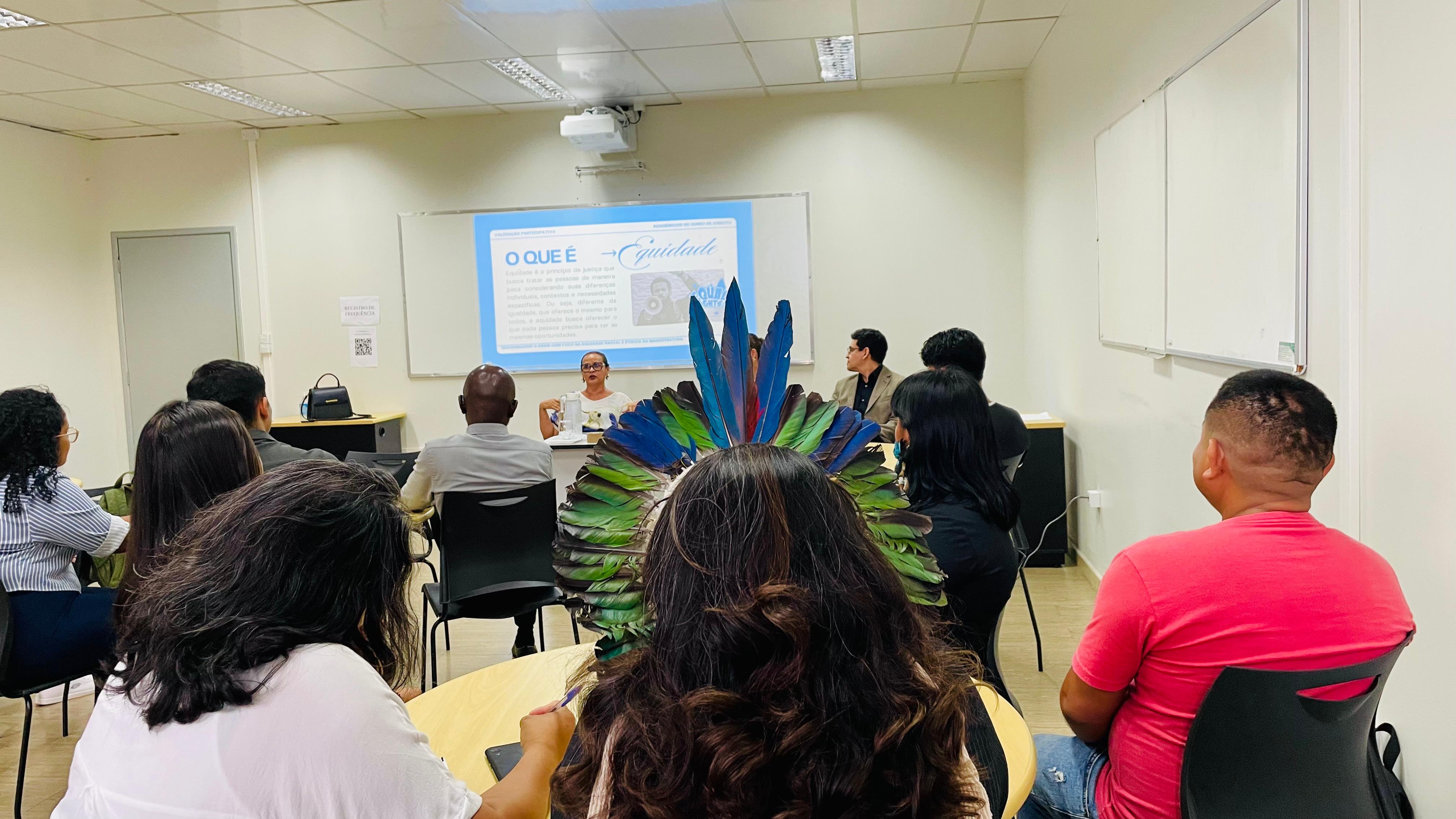 Imagem colorida mostra a reunião com estudantes sobre equidade racial no Exame Nacional da Magistratura em uma sala de aula. Ao centro da imagem, destaca-se o cocar de penas coloridas de uma das participantes. No fundo, uma mulher fala diante de um slide projetado com o título “O que é equidade”, enquanto os demais participantes a escutam.