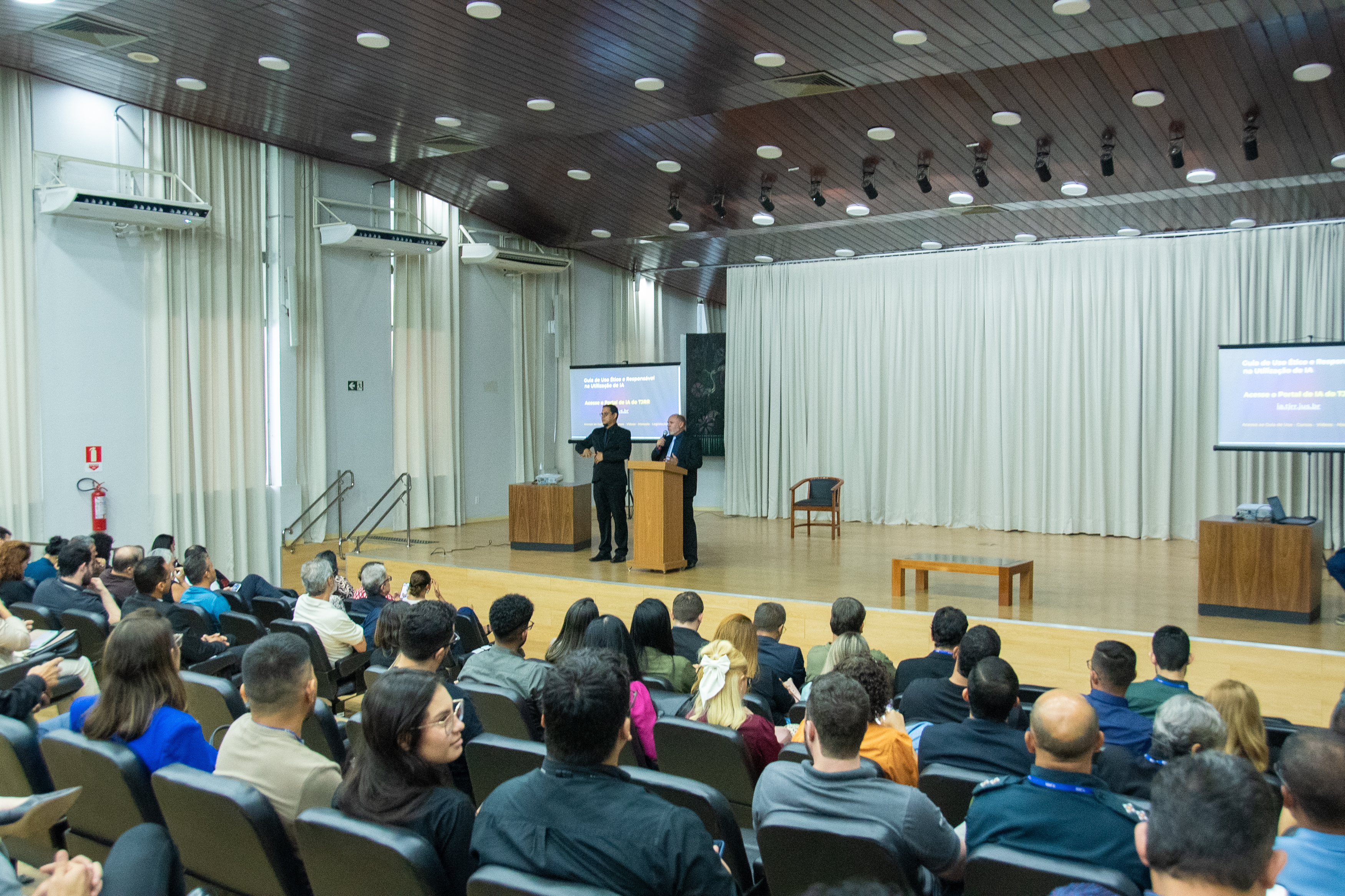 A imagem colorida mostra um auditório durante um workshop. Em primeiro plano, a parte de trás da plateia é visível, com várias pessoas sentadas em cadeiras estofadas escuras, voltadas para o palco. No palco, que é de madeira clara, uma mulher e um homem estão de pé. O homem, à esquerda, o interprete de Libras, enquanto a mulher, à direita, está em frente a um púlpito. Há dois púlpitos de madeira no palco, um ao lado do outro, e um banco de madeira mais baixo entre eles. Ao fundo do palco, cortinas claras cobrem a parede.  Texto: Mairon Compagnon - Jornalista 