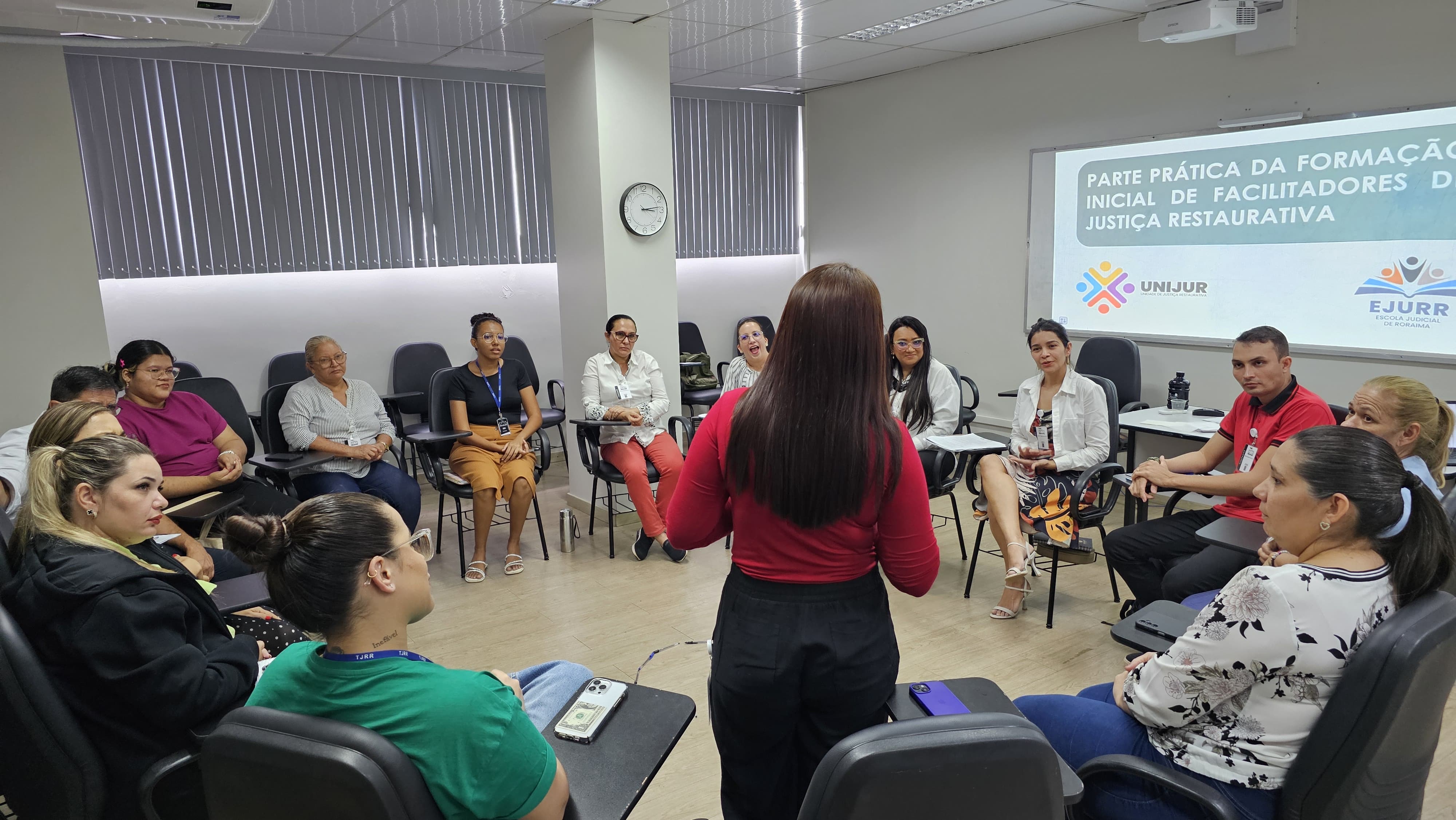 foto colorida de pessoas sentadas em círculos e a frente há uma imagem projetada da apresentação. foto colorida de pessoas sentadas em círculos, uma está em pé a frente há uma imagem projetada da apresentação.