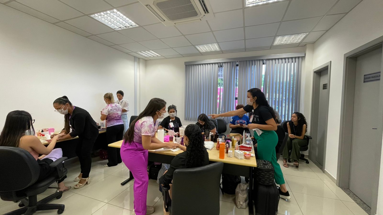Imagem colorida mostra uma sala com várias mulheres realizando atendimentos. Algumas estão sentadas recebendo cuidados nas mãos, enquanto outras trabalham com produtos de beleza dispostos sobre a mesa. As profissionais utilizam máscaras de proteção.