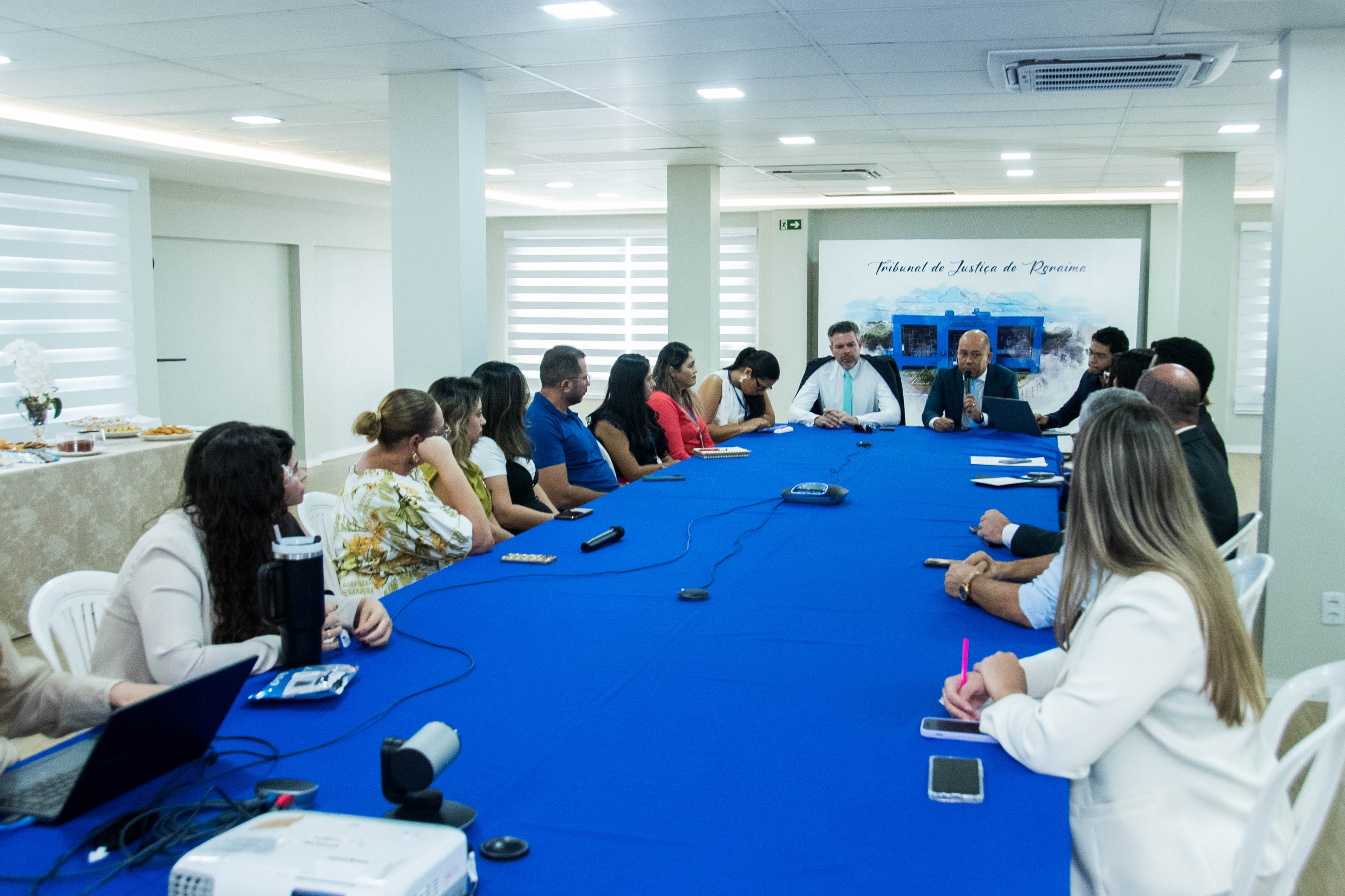 Foto colorida mostra uma reunião de cerca de 15 pessoas sentadas ao redor de uma grande mesa azul. Foto colorida mostra uma reunião de cerca de 15 pessoas sentadas ao redor de uma grande mesa azul.