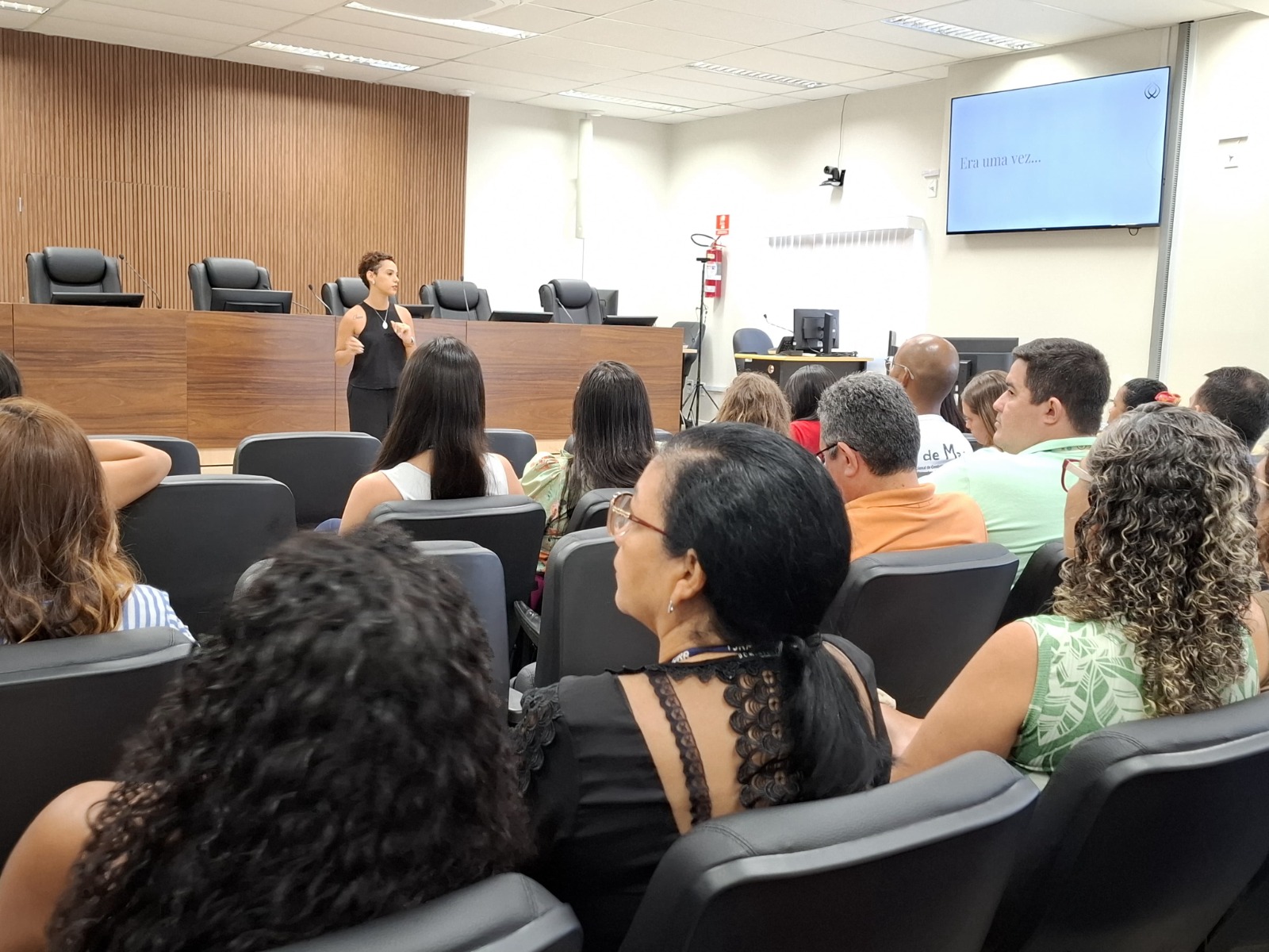 Imagem colorida mostra uma palestra em andamento em um auditório. Uma mulher está em pé, em frente ao público, falando e gesticulando. Ao fundo, há uma bancada com cadeiras de juiz e um painel de madeira. O público está sentado e atento, assistindo. Imagem colorida mostra uma palestra em andamento em um auditório. Uma mulher está em pé, em frente ao público, falando e gesticulando. Ao fundo, há uma bancada com cadeiras de juiz e um painel de madeira. O público está sentado e atento, assistindo.