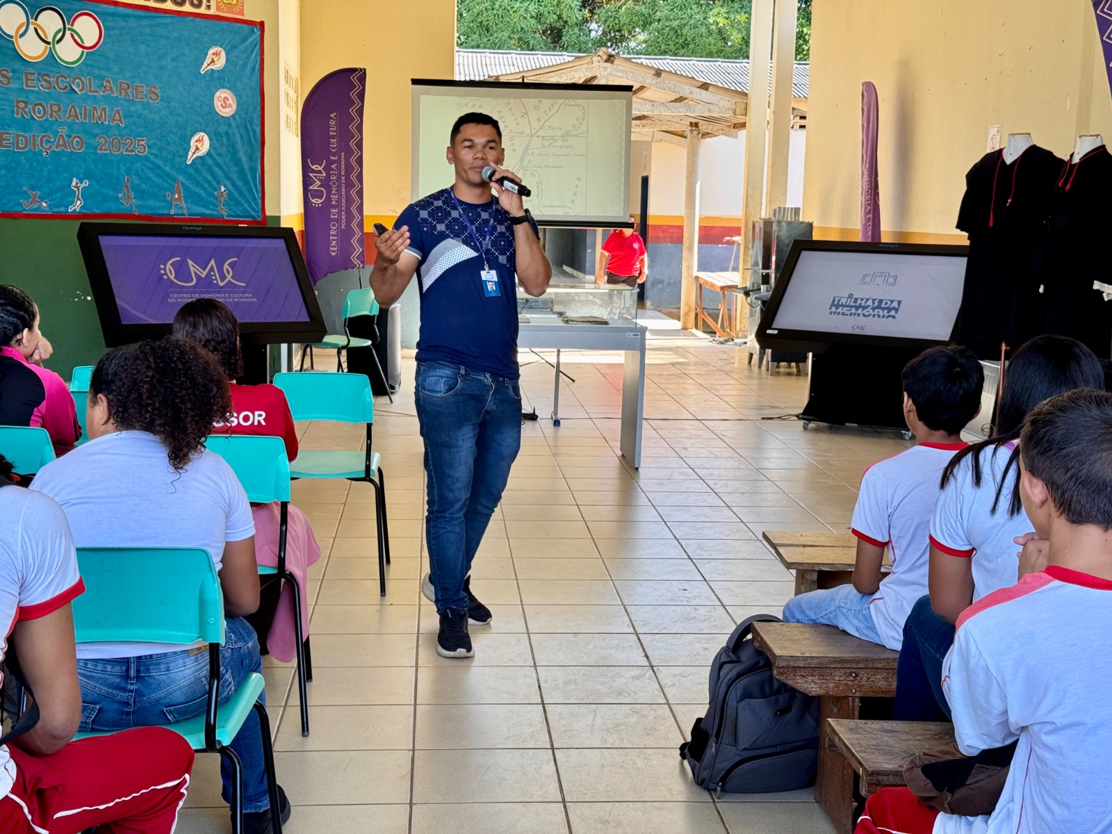 A imagem colorida mostra um homem em pé, no centro, vestindo uma camiseta azul e calça jeans, segurando um microfone e gesticulando enquanto fala para um grupo de jovens sentados em cadeiras e bancos à sua frente. Ao fundo, há um telão com projeção, e à direita e à esquerda, painéis digitais. O ambiente é um espaço aberto, parcialmente coberto, que parece ser uma área de eventos ou pátio escolar, com árvores e construções ao fundo. No lado esquerdo, um banner azul com o logotipo olímpico e o texto "Jogos Escolares de Roraima Edição 2025" é visível. A imagem colorida mostra um homem em pé, no centro, vestindo uma camiseta azul e calça jeans, segurando um microfone e gesticulando enquanto fala para um grupo de jovens sentados em cadeiras e bancos à sua frente. Ao fundo, há um telão com projeção, e à direita e à esquerda, painéis digitais. O ambiente é um espaço aberto, parcialmente coberto, que parece ser uma área de eventos ou pátio escolar, com árvores e construções ao fundo. No lado esquerdo, um banner azul com o logotipo olímpico e o texto "Jogos Escolares de Roraima Edição 2025" é visível.