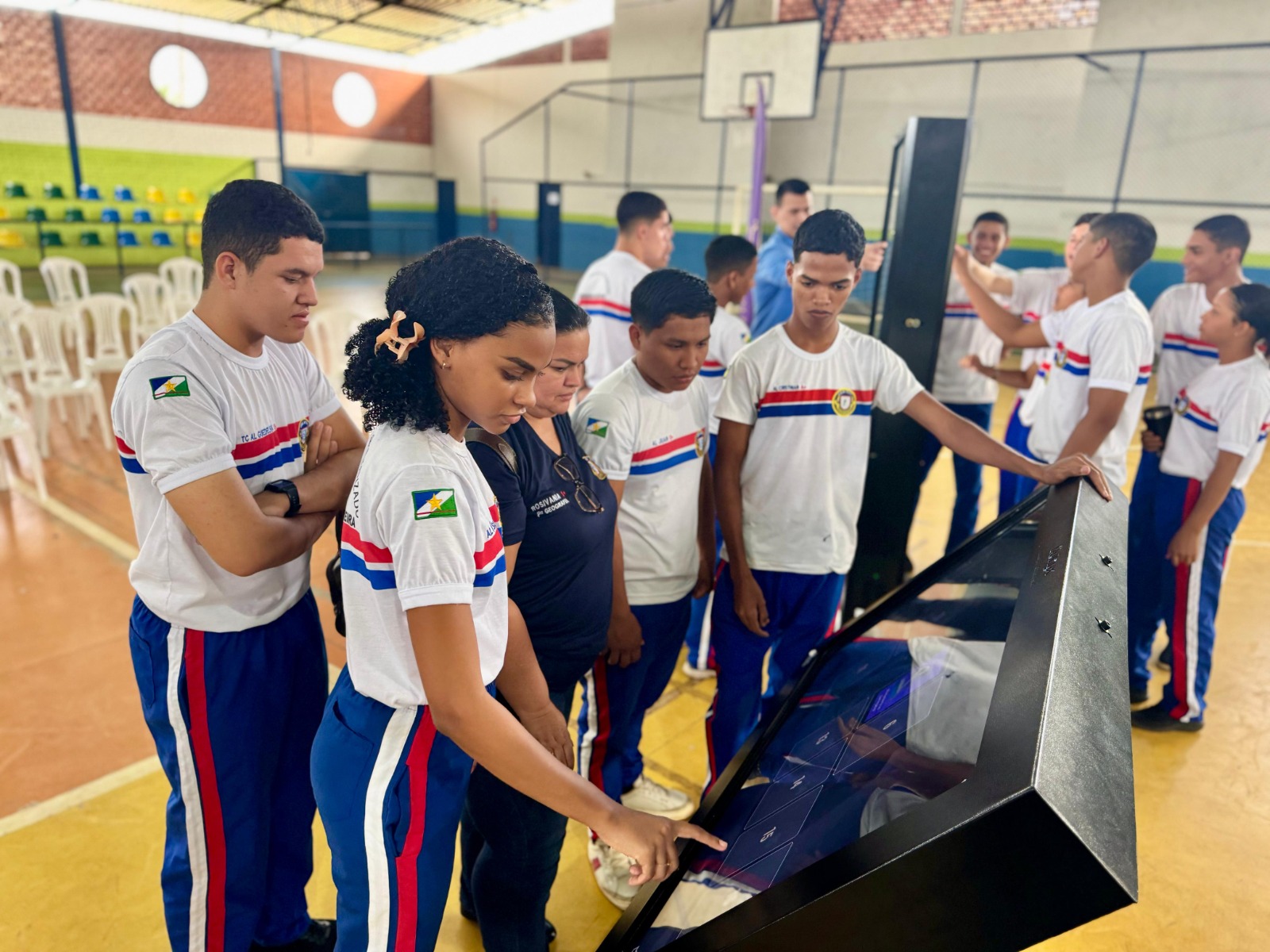 A imagem colorida mostra um grupo de jovens, a maioria vestindo uniformes de camiseta branca e calça azul com listras laterais vermelhas e brancas, reunidos ao redor de uma tela interativa inclinada, de cor escura. Uma jovem no centro da imagem está apontando para a tela, enquanto os outros a observam. O ambiente é interno, possivelmente um ginásio ou salão escolar, com o chão em tons de laranja e algumas cadeiras brancas ao fundo. A imagem colorida mostra um grupo de jovens, a maioria vestindo uniformes de camiseta branca e calça azul com listras laterais vermelhas e brancas, reunidos ao redor de uma tela interativa inclinada, de cor escura. Uma jovem no centro da imagem está apontando para a tela, enquanto os outros a observam. O ambiente é interno, possivelmente um ginásio ou salão escolar, com o chão em tons de laranja e algumas cadeiras brancas ao fundo.