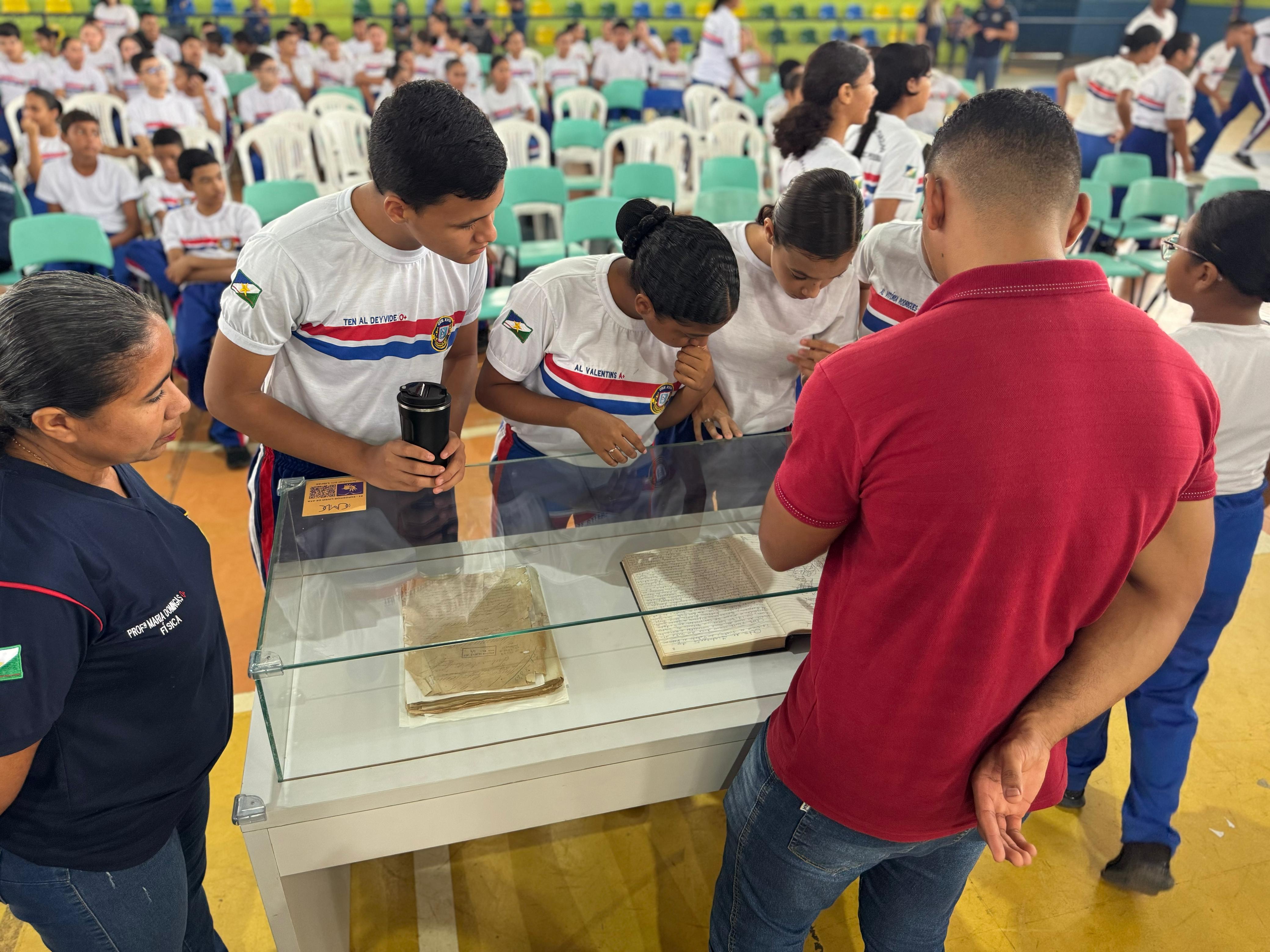 A imagem colorida mostra cinco pessoas, possivelmente estudantes e educadores, em volta de uma vitrine de vidro que exibe dois objetos antigos, provavelmente livros ou documentos. O ambiente ao redor parece ser um ginásio ou auditório, com muitas cadeiras brancas e outras pessoas sentadas ao fundo. As pessoas em primeiro plano estão atentas aos objetos na vitrine. A imagem colorida mostra cinco pessoas, possivelmente estudantes e educadores, em volta de uma vitrine de vidro que exibe dois objetos antigos, provavelmente livros ou documentos. O ambiente ao redor parece ser um ginásio ou auditório, com muitas cadeiras brancas e outras pessoas sentadas ao fundo. As pessoas em primeiro plano estão atentas aos objetos na vitrine.