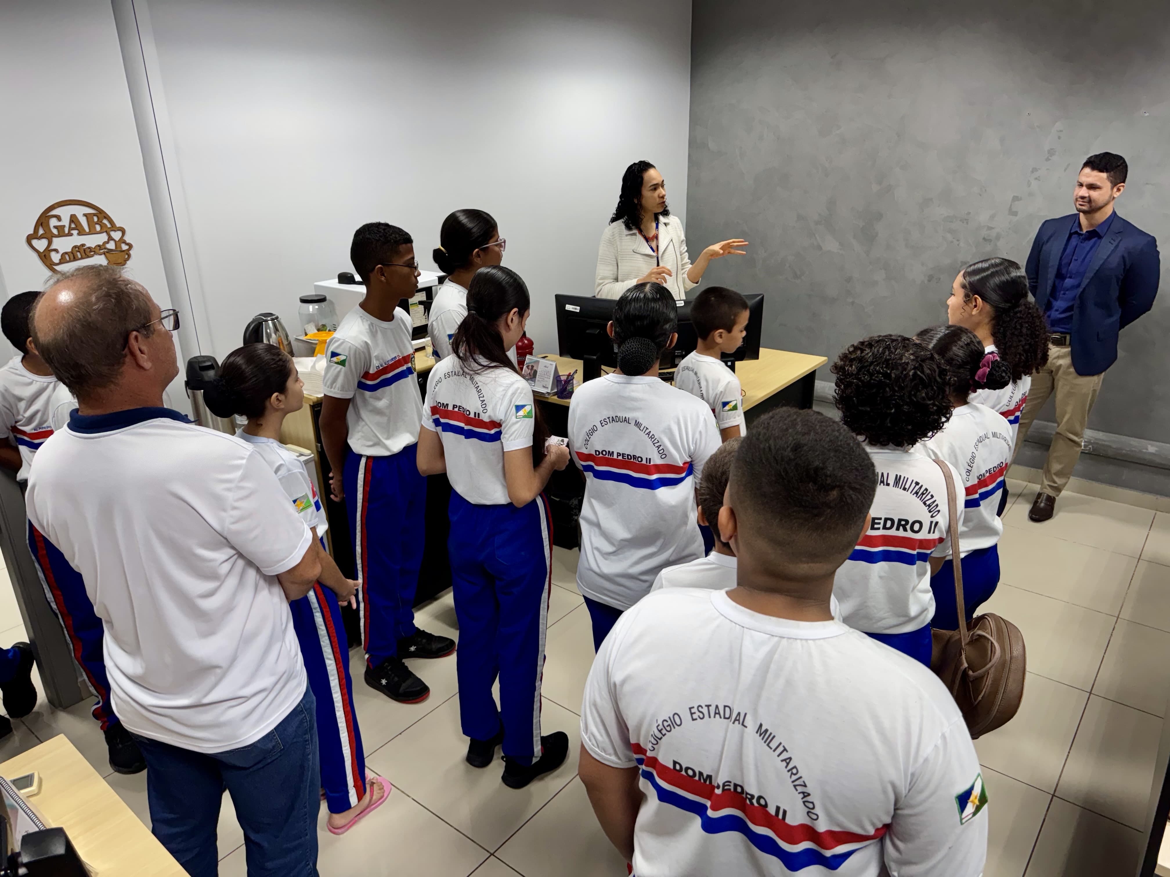 Imagem colorida mostra um grupo de estudantes do Colégio Dom Pedro II em uma sala. Eles estão de pé, ouvindo atentamente uma mulher que fala próximo a uma mesa com computador. O juiz Cleber Gonçalves também acompanha a atividade, posicionado à frente do grupo, próximo à parede.