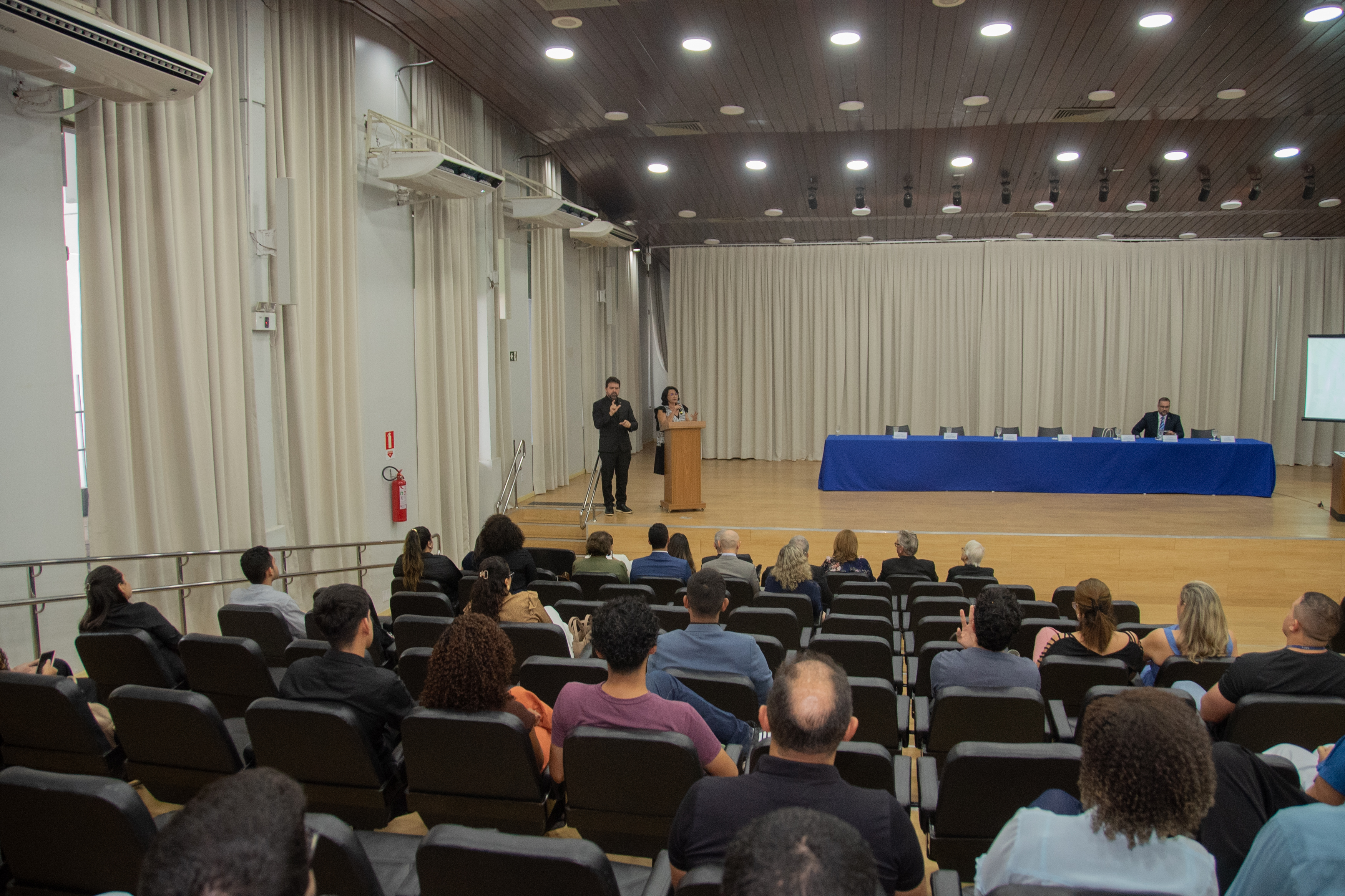 A imagem mostra um auditório com plateia vista de trás, atenta a um evento no palco. Duas pessoas estão em pé no centro do palco, uma delas falando ao microfone em um púlpito. Há uma mesa com um homem sentado, cortinas claras ao fundo, telão parcialmente visível e cadeiras escuras organizadas em fileiras com corredor central. A imagem mostra um auditório com plateia vista de trás, atenta a um evento no palco. Duas pessoas estão em pé no centro do palco, uma delas falando ao microfone em um púlpito. Há uma mesa com um homem sentado, cortinas claras ao fundo, telão parcialmente visível e cadeiras escuras organizadas em fileiras com corredor central.