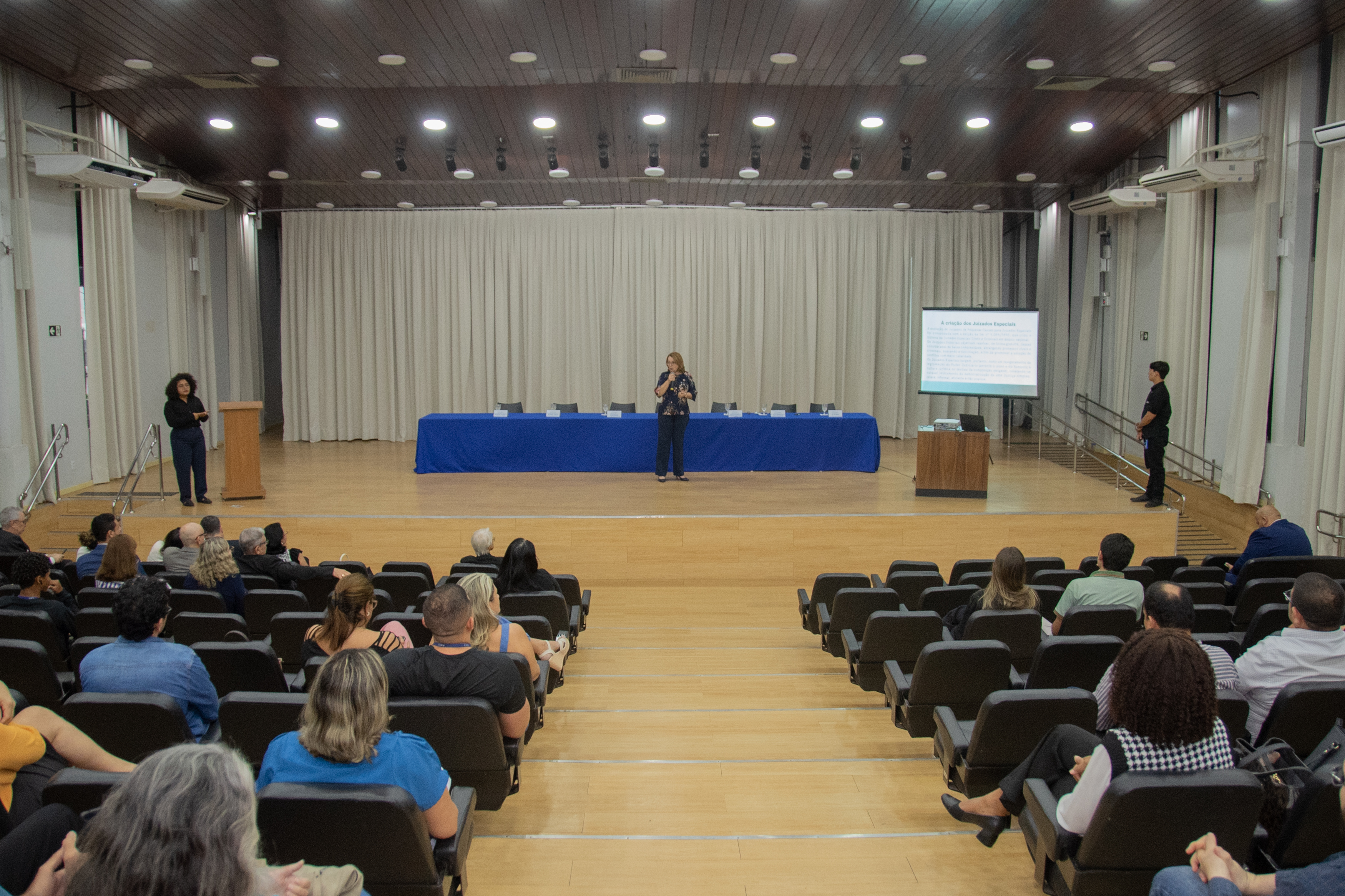 A imagem mostra um auditório com plateia atenta e Desembargadora Elaine Bianchi está no palco fazendo uma apresentação. Ela está em pé, gesticulando, enquanto ao fundo há uma mesa coberta, um telão com slides projetados e pessoas em pé nas laterais do palco. O ambiente é iluminado e transmite o clima de uma palestra ou evento formal. A imagem mostra um auditório com plateia atenta e Desembargadora Elaine Bianchi está no palco fazendo uma apresentação. Ela está em pé, gesticulando, enquanto ao fundo há uma mesa coberta, um telão com slides projetados e pessoas em pé nas laterais do palco. O ambiente é iluminado e transmite o clima de uma palestra ou evento formal.