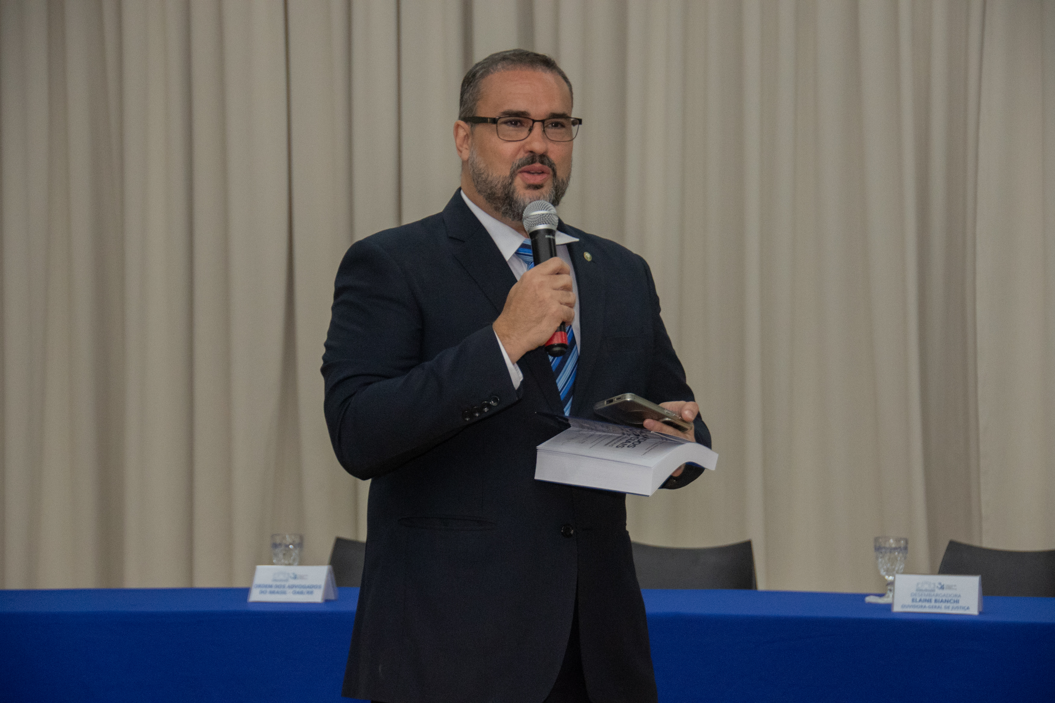 A imagem mostra o juiz Felippe Borring Rocha. Ele veste terno azul, segura microfone, livro e celular. Está em frente a uma mesa com toalha azul, com copos e cartões. Ao fundo, há uma cortina clara e cadeiras parcialmente visíveis. A imagem mostra o juiz Felippe Borring Rocha. Ele veste terno azul, segura microfone, livro e celular. Está em frente a uma mesa com toalha azul, com copos e cartões. Ao fundo, há uma cortina clara e cadeiras parcialmente visíveis.