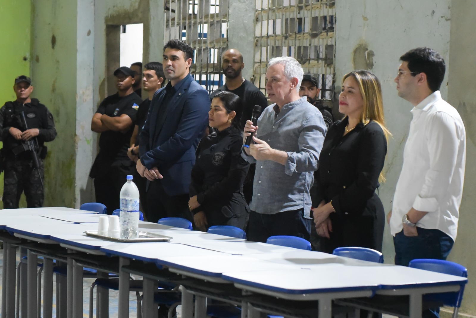Imagem colorida mostra um grupo de pessoas em pé, incluindo homens e mulheres, alguns em roupas sociais escuras e claras, e outros em uniformes. Ao centro, um homem decabelo grisalho com uma camisa social cinza clara está segurando um microfone e falando. À direita, há uma mesa comprida com garrafas de água e copos. Ao fundo, uma grade na janela. Imagem colorida mostra um grupo de pessoas em pé, incluindo homens e mulheres, alguns em roupas sociais escuras e claras, e outros em uniformes. Ao centro, um homem decabelo grisalho com uma camisa social cinza clara está segurando um microfone e falando. À direita, há uma mesa comprida com garrafas de água e copos. Ao fundo, uma grade na janela.