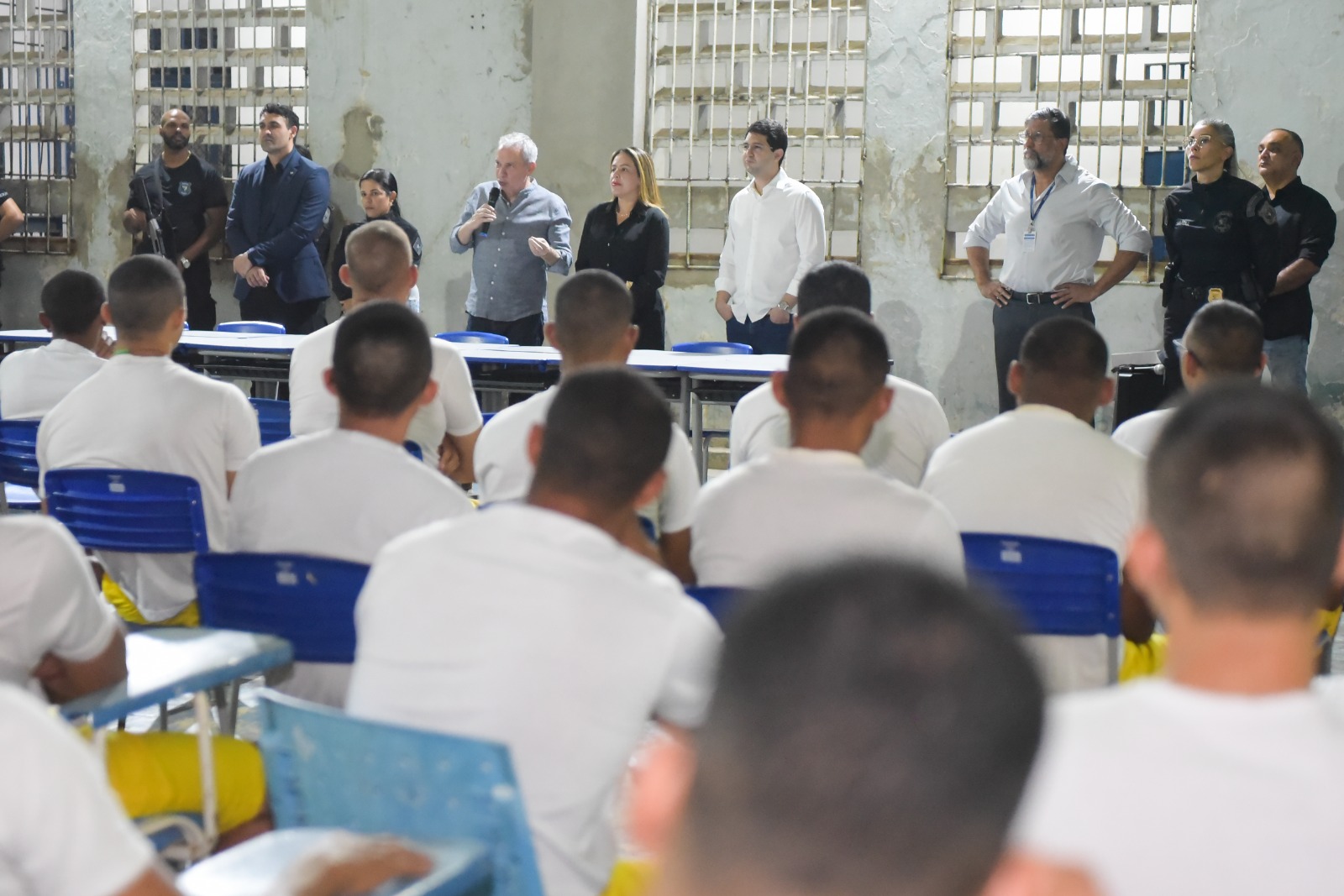 Imagem colorida mostra uma reunião ou evento em um ambiente interno, com várias pessoas sentadas em mesas escolares azuis e brancas, voltadas para uma pequena plataforma. Na plataforma, um grupo de indivíduos em pé, incluindo homens e mulheres, alguns vestindo camisas sociais claras e outros uniformes escuros, estão presentes. Um homem de cabelo grisalho no centro da plataforma está falando em um microfone. Imagem colorida mostra uma reunião ou evento em um ambiente interno, com várias pessoas sentadas em mesas escolares azuis e brancas, voltadas para uma pequena plataforma. Na plataforma, um grupo de indivíduos em pé, incluindo homens e mulheres, alguns vestindo camisas sociais claras e outros uniformes escuros, estão presentes. Um homem de cabelo grisalho no centro da plataforma está falando em um microfone.
