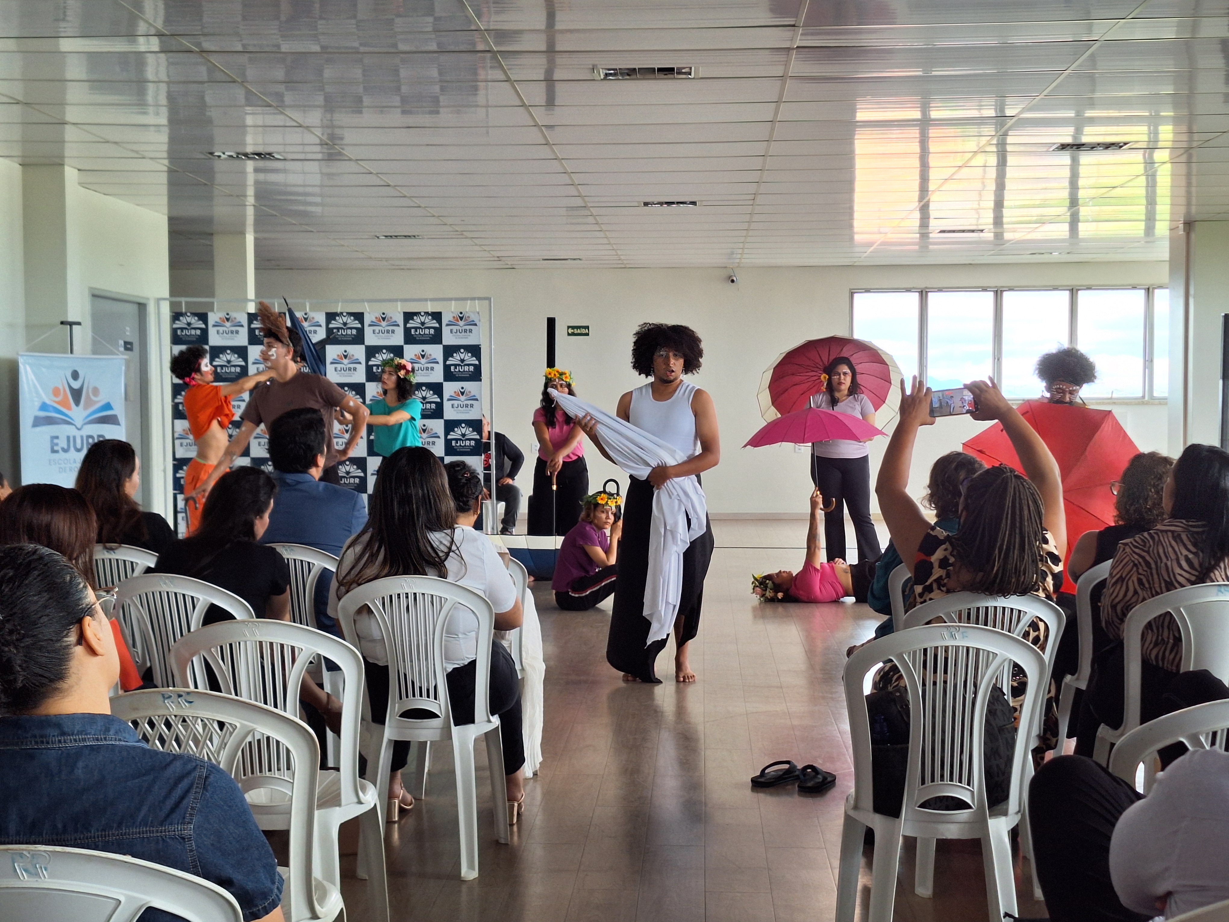 Grupo de performers ou estudantes em fila em um salão, alguns em trajes de performance, enquanto um indivíduo de preto interage com a pessoa da extrema direita. Grupo de performers ou estudantes em fila em um salão, alguns em trajes de performance, enquanto um indivíduo de preto interage com a pessoa da extrema direita.